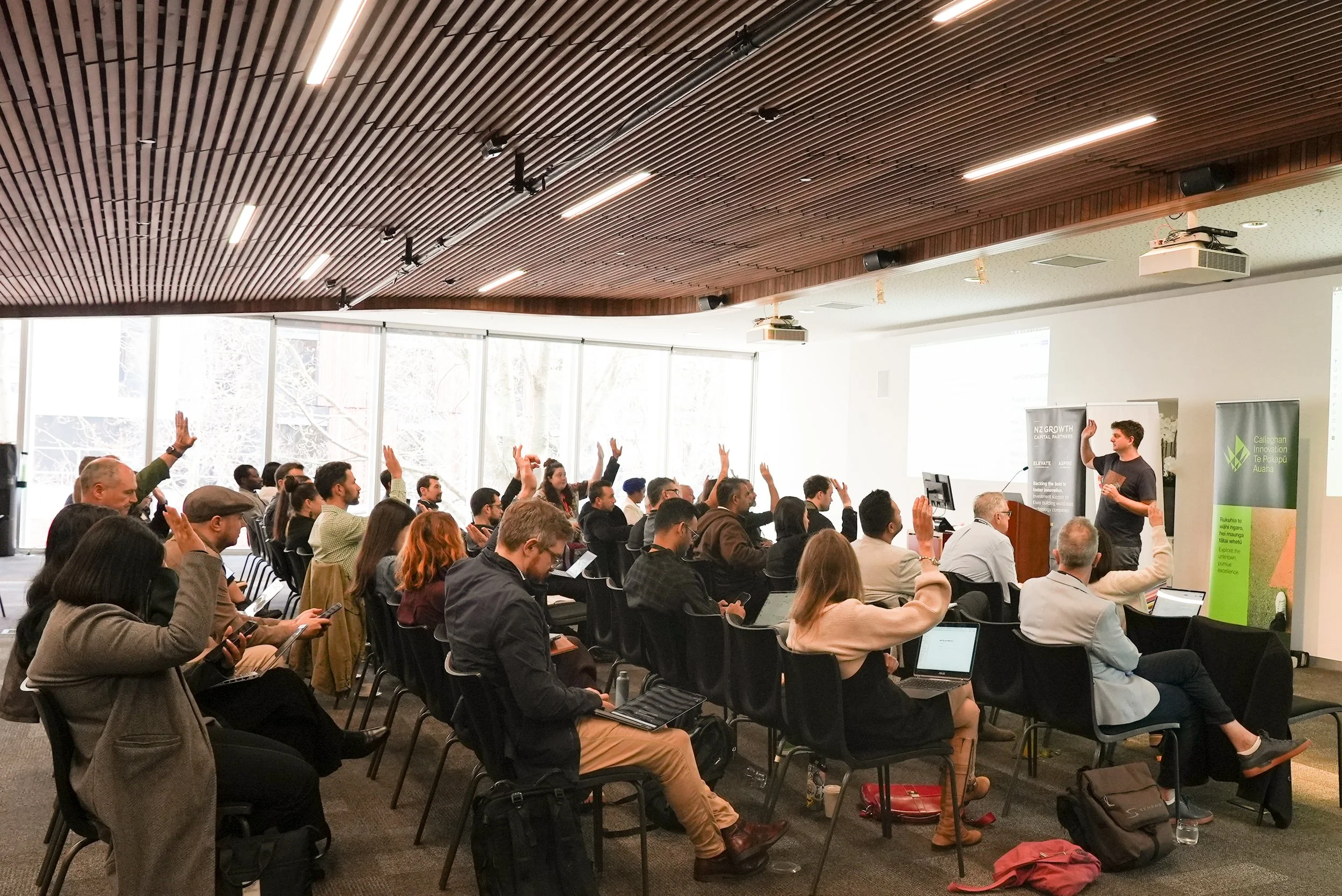 A conference or seminar with an audience of diverse adults seated in rows, some raising their hands, facing a speaker at the front of the room. The room has large windows, a wooden ceiling, and presentation screens.