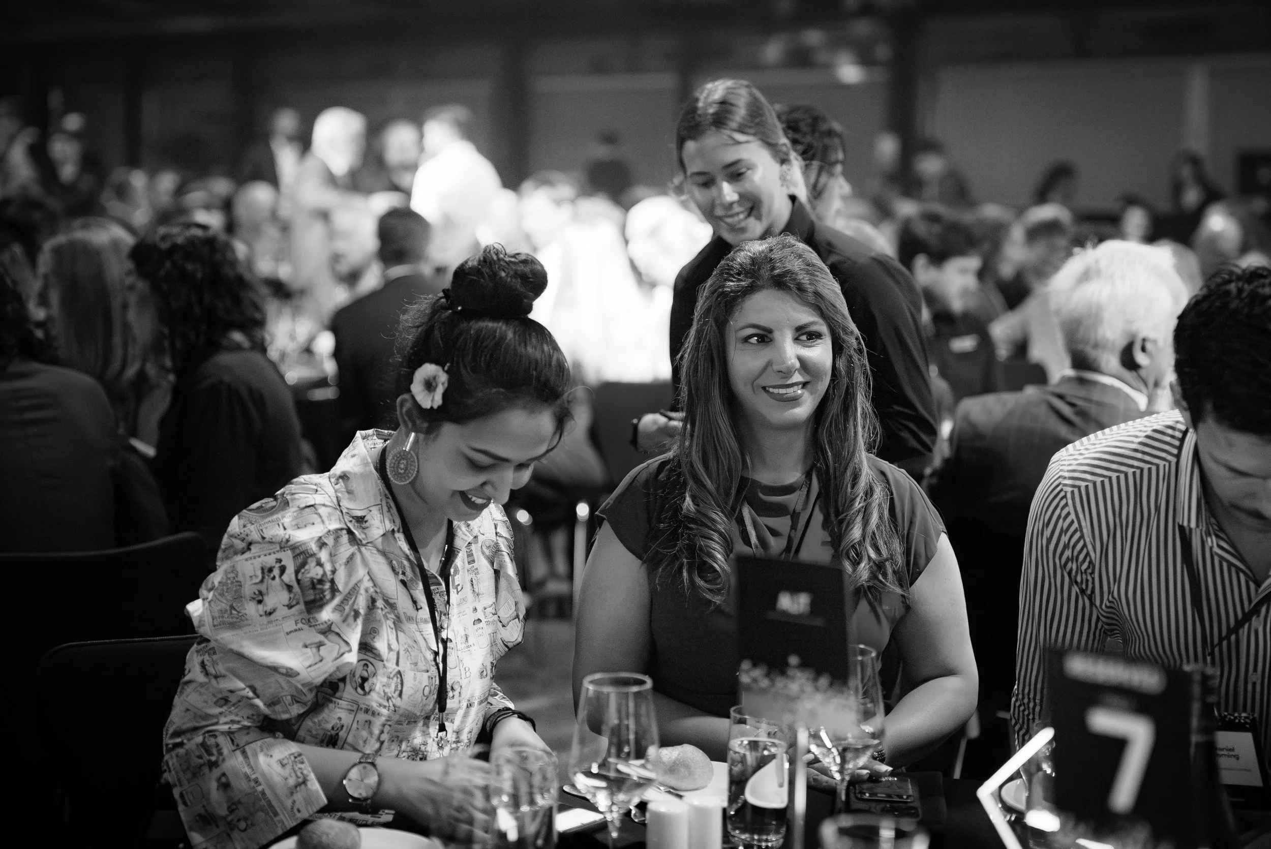 A group of diverse women at a formal event, sitting at a table with glasses and tableware, engaged in conversation and smiling.