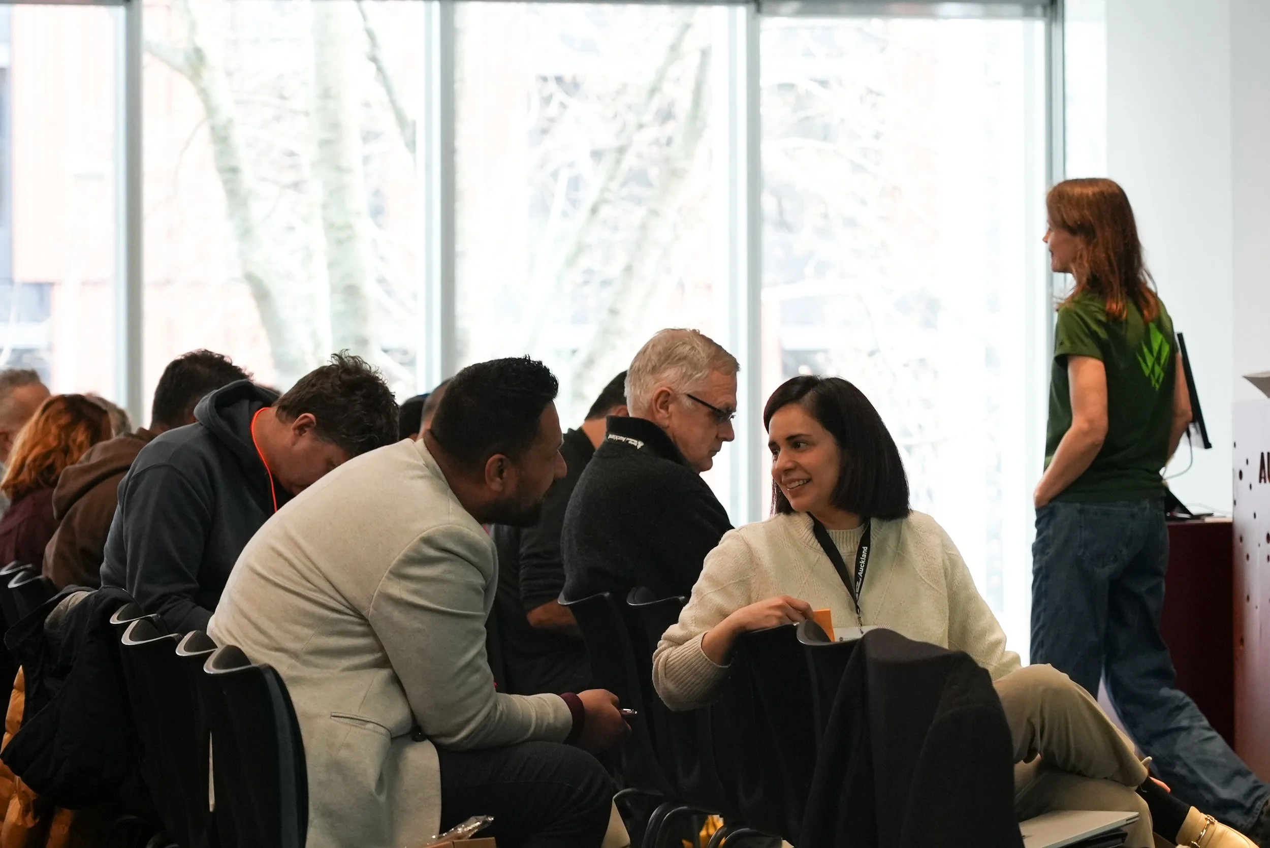People sitting in a conference room near large windows, engaging in conversation and listening to a speaker.
