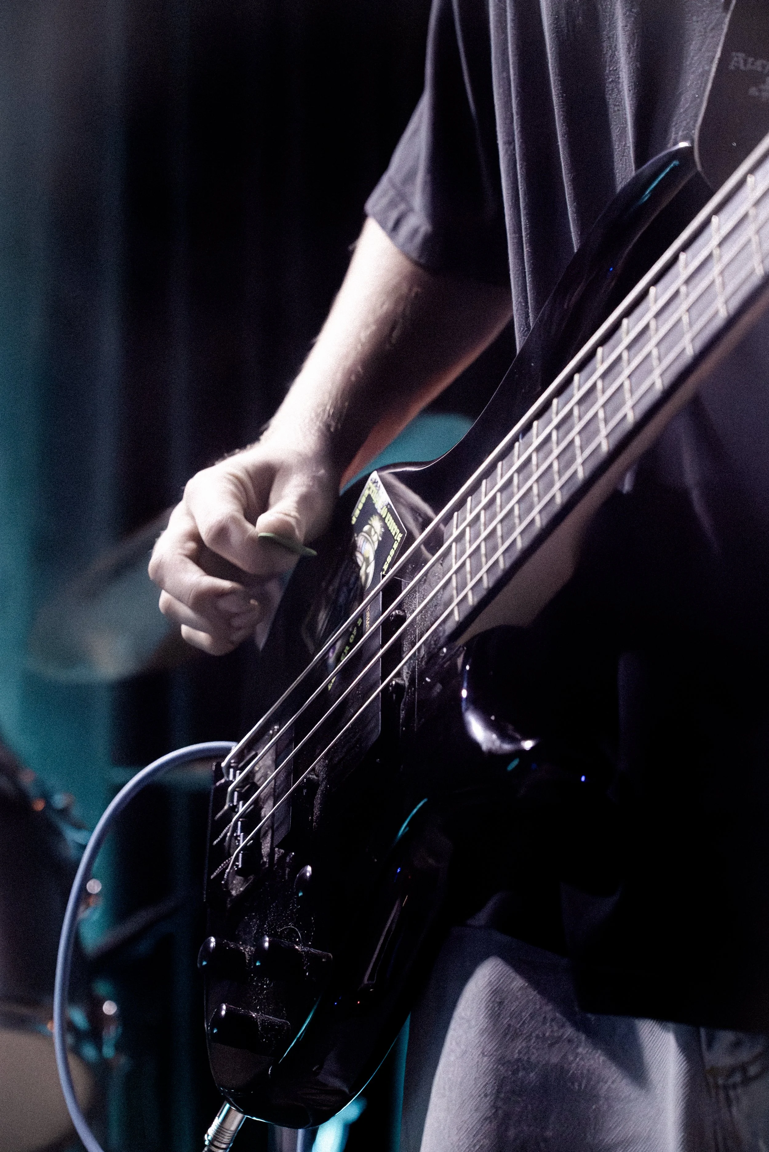 Close-up of a person playing a black electric bass guitar, focusing on their hand on the strings and part of their torso, with a dark background.