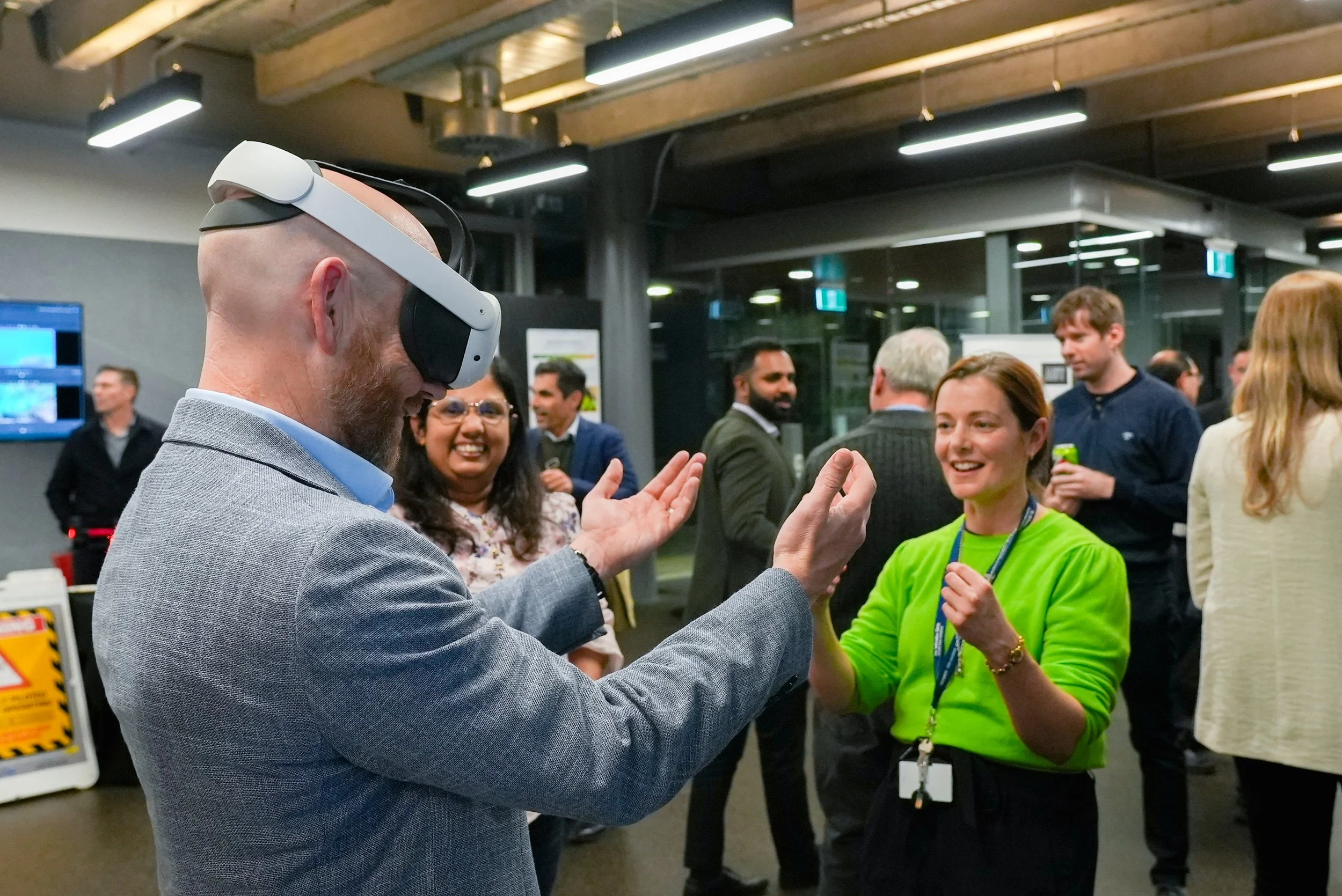 A man in a gray blazer wearing a virtual reality headset interacts with a woman in a green shirt at a networking event, with other people socializing in the background.