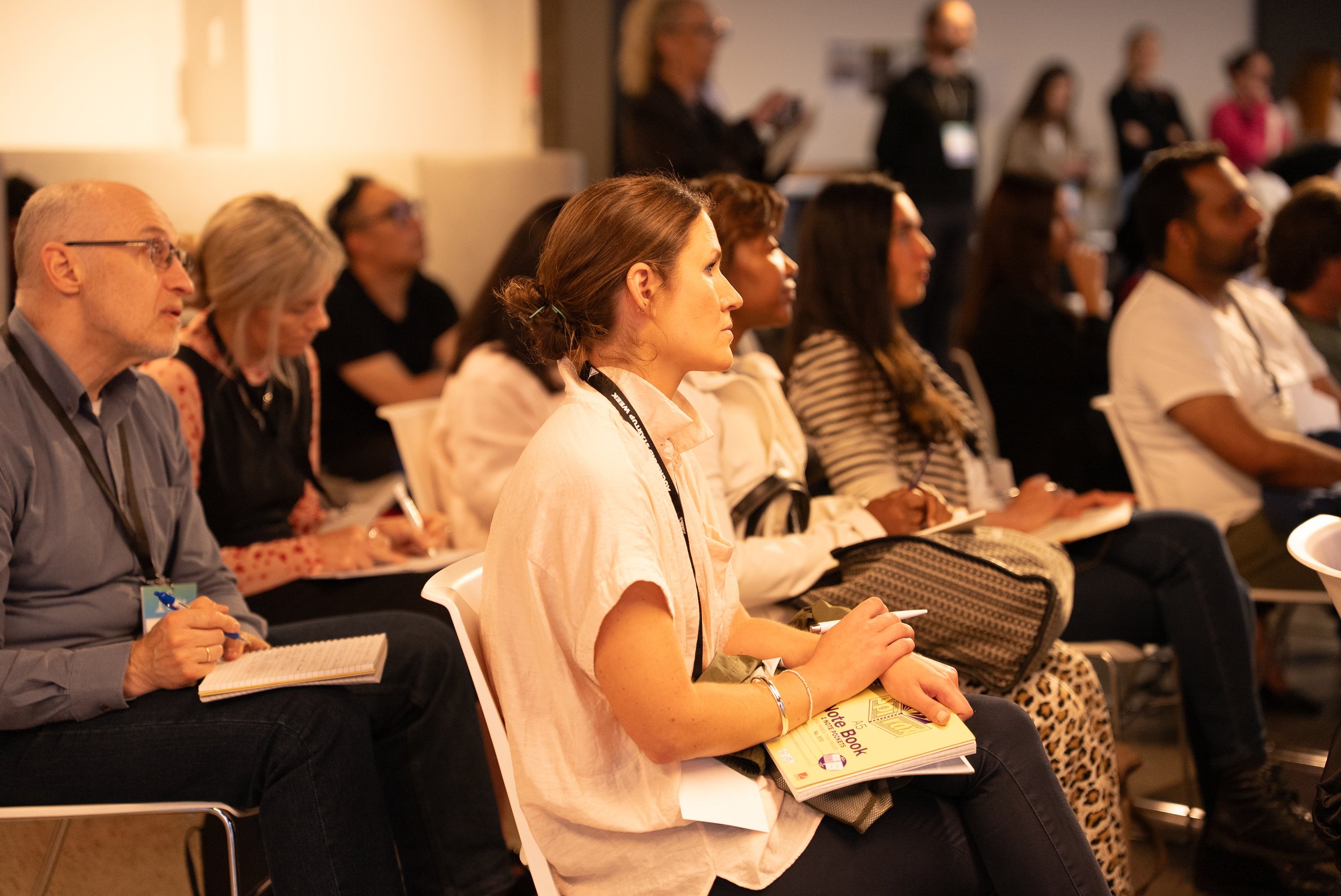 Audience of men and women sitting attentively at a conference, listening to a speaker, some taking notes, with a woman in the foreground holding a yellow notebook.