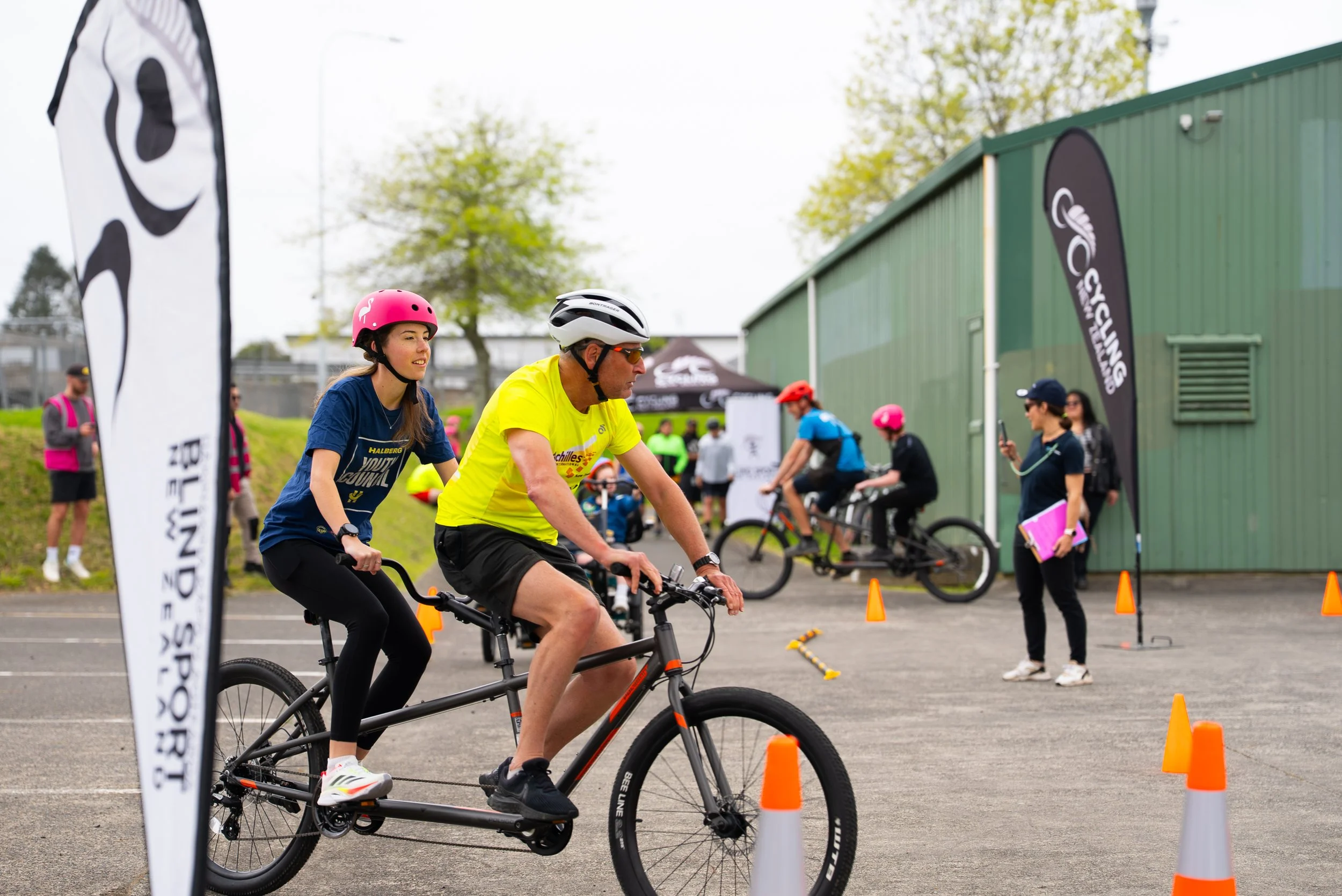A group of people participating in a bicycle training or event, with two riders in the foreground, a young woman and an older man, both wearing helmets. The scene is set outdoors near a green building, with flags and cones marking the course, and several other riders and spectators in the background.