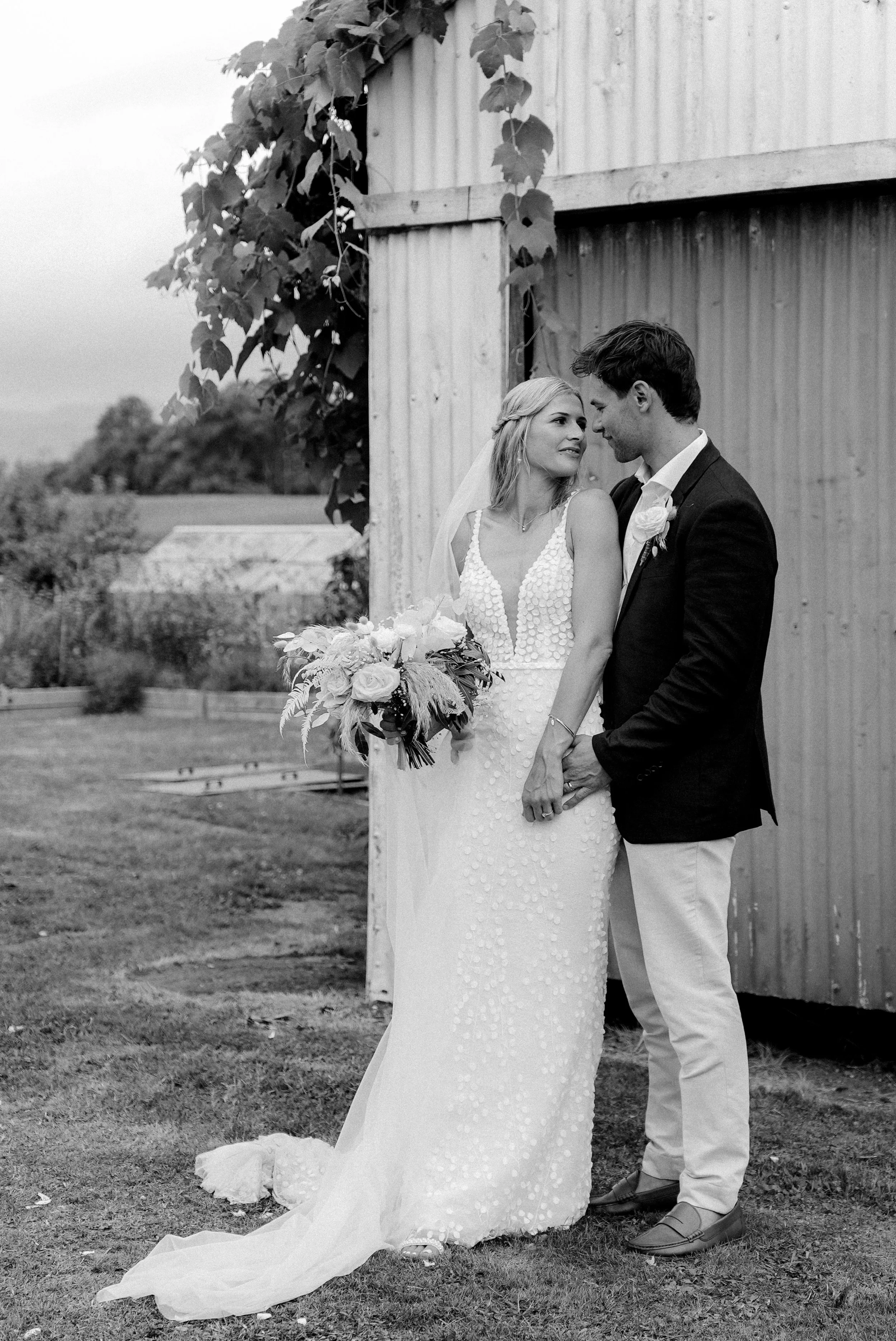 Black and white photo of a bride and groom standing outdoors, holding hands, close to a rustic barn with vines growing on it. The bride is in a sleeveless, beaded wedding gown holding a bouquet, and the groom is in a dark blazer, light trousers, and dress shoes.