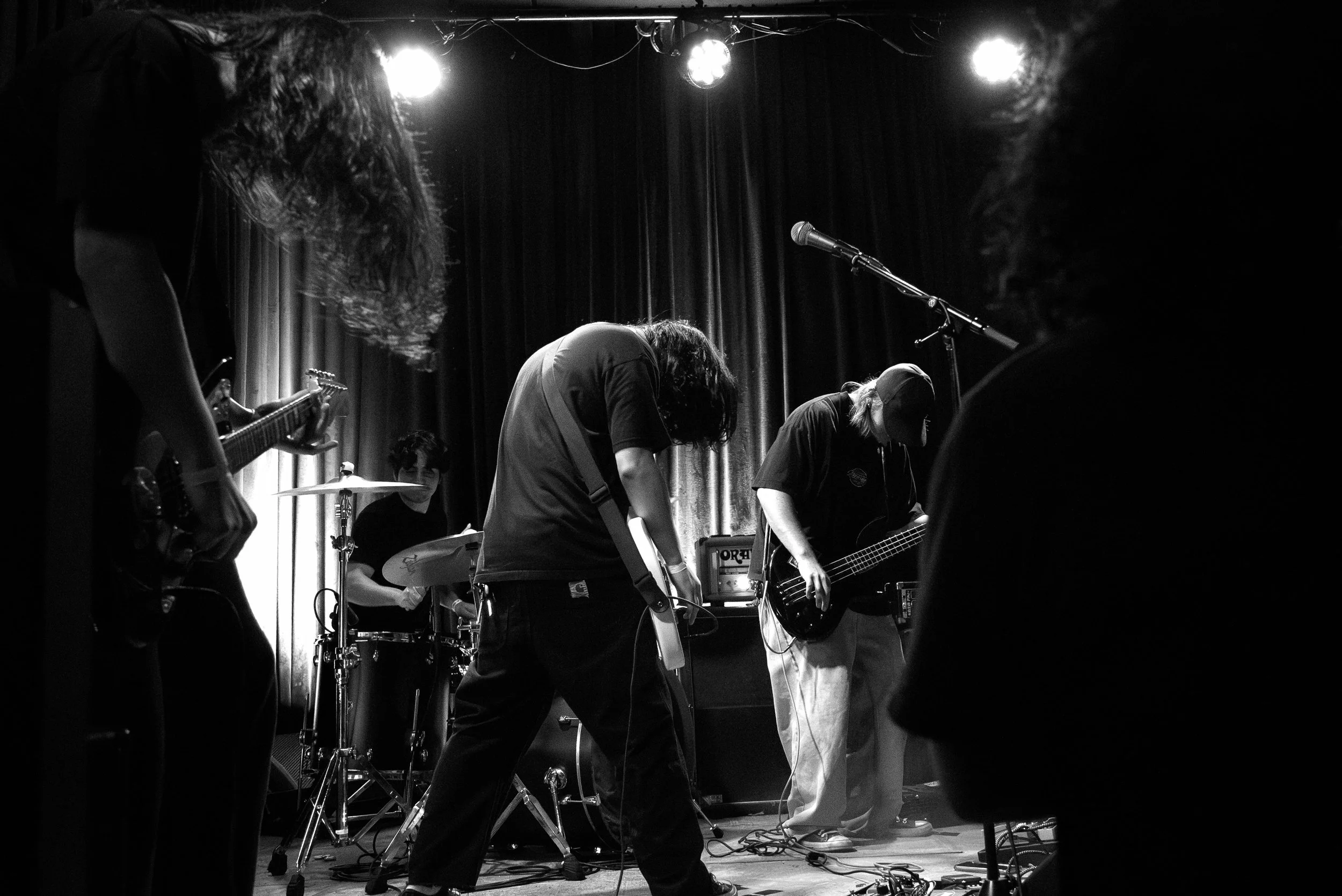 A black and white photo of a band performing on stage, with multiple band members playing guitars, drums, and bass, under stage lights with a curtain backdrop.