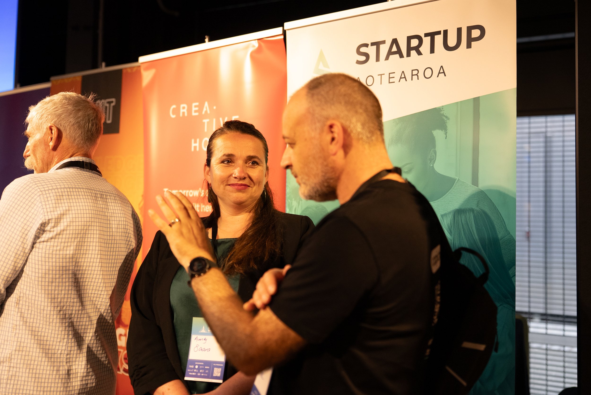 Three people are engaged in conversation at a professional event. Two are visible in the foreground, with one woman smiling and another man gesturing with his hand. A third person is partially visible on the left. Behind them are colorful banners with text related to startups and innovation.