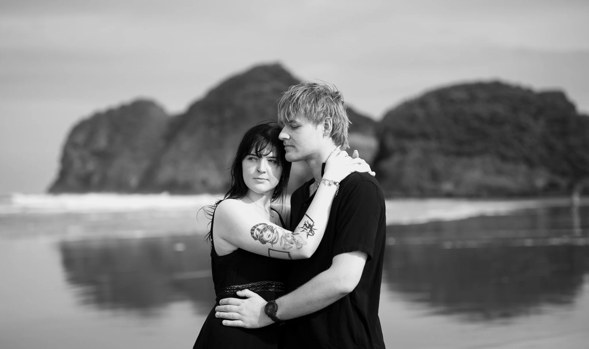 A black-and-white photo of a couple embracing on a beach with rock formations in the background. The woman has tattoos and dark hair, and the man has light hair and is wearing a watch.
