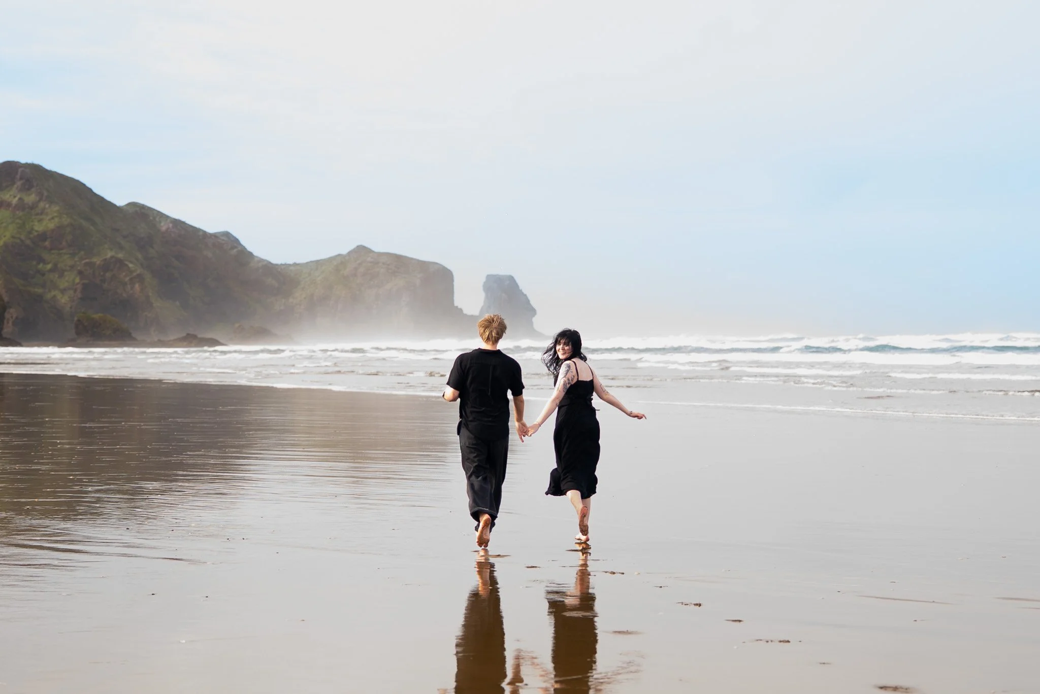 Two people holding hands, walking on the beach near the ocean with cliffs in the background.