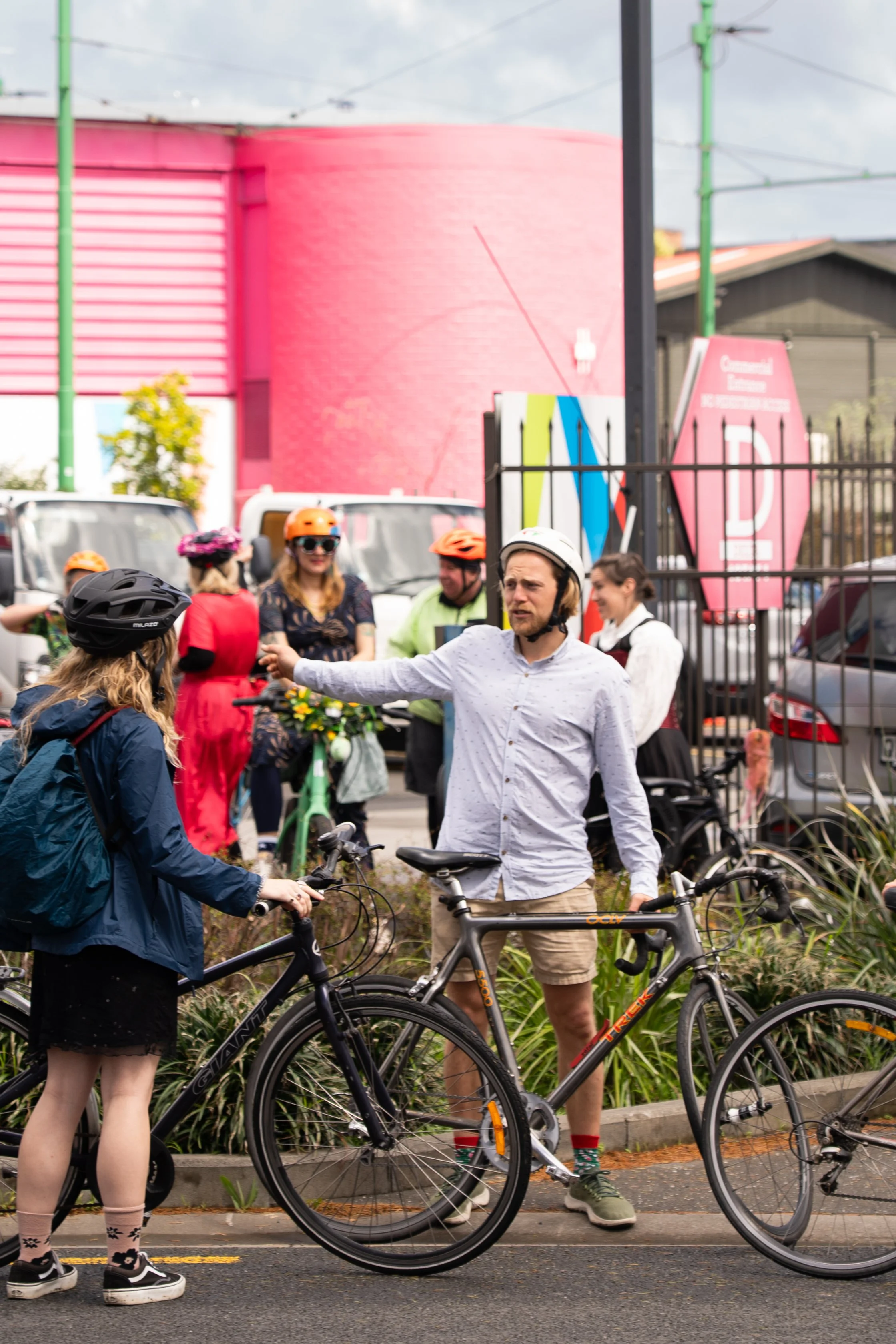 People with bicycles on a city street, some wearing helmets, with a colorful pink and green building in the background.