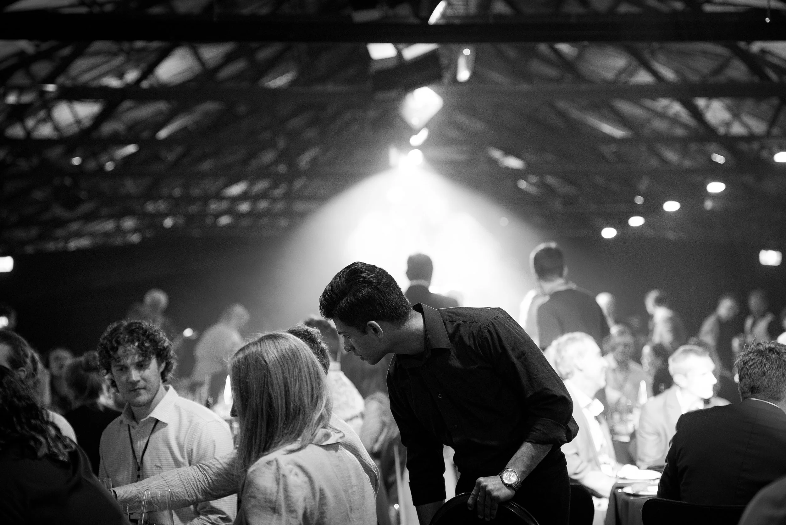 Black and white photo of a banquet or event with people sitting at tables, some engaged in conversation, and a young man in a black shirt leaning over a table.