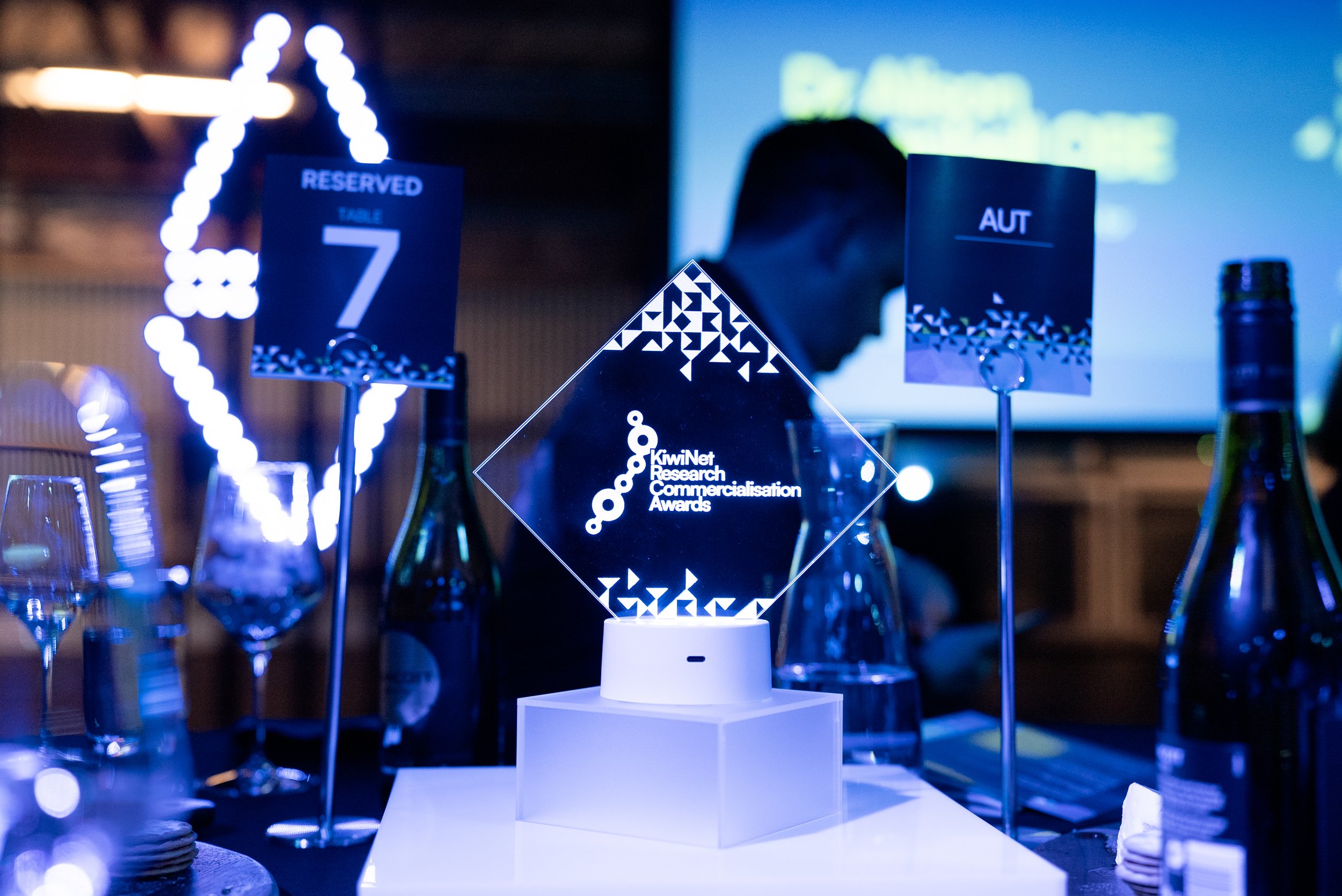 Glass award with geometric designs and the text 'KiwiNet Research Commercialisation Awards' displayed on a table at an event, surrounded by table markers, wine bottles, and glasses.