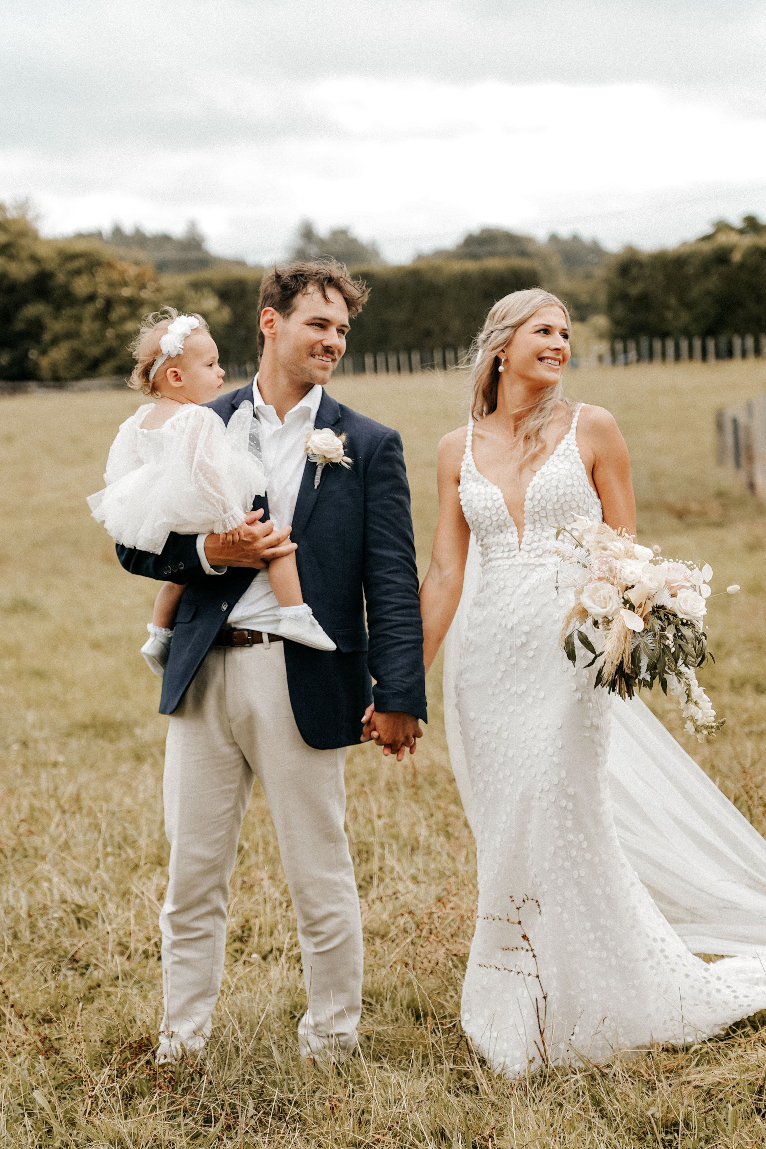 A newlywed couple holding hands, with a man carrying a young girl in a white dress, in a grassy outdoor field during daytime.