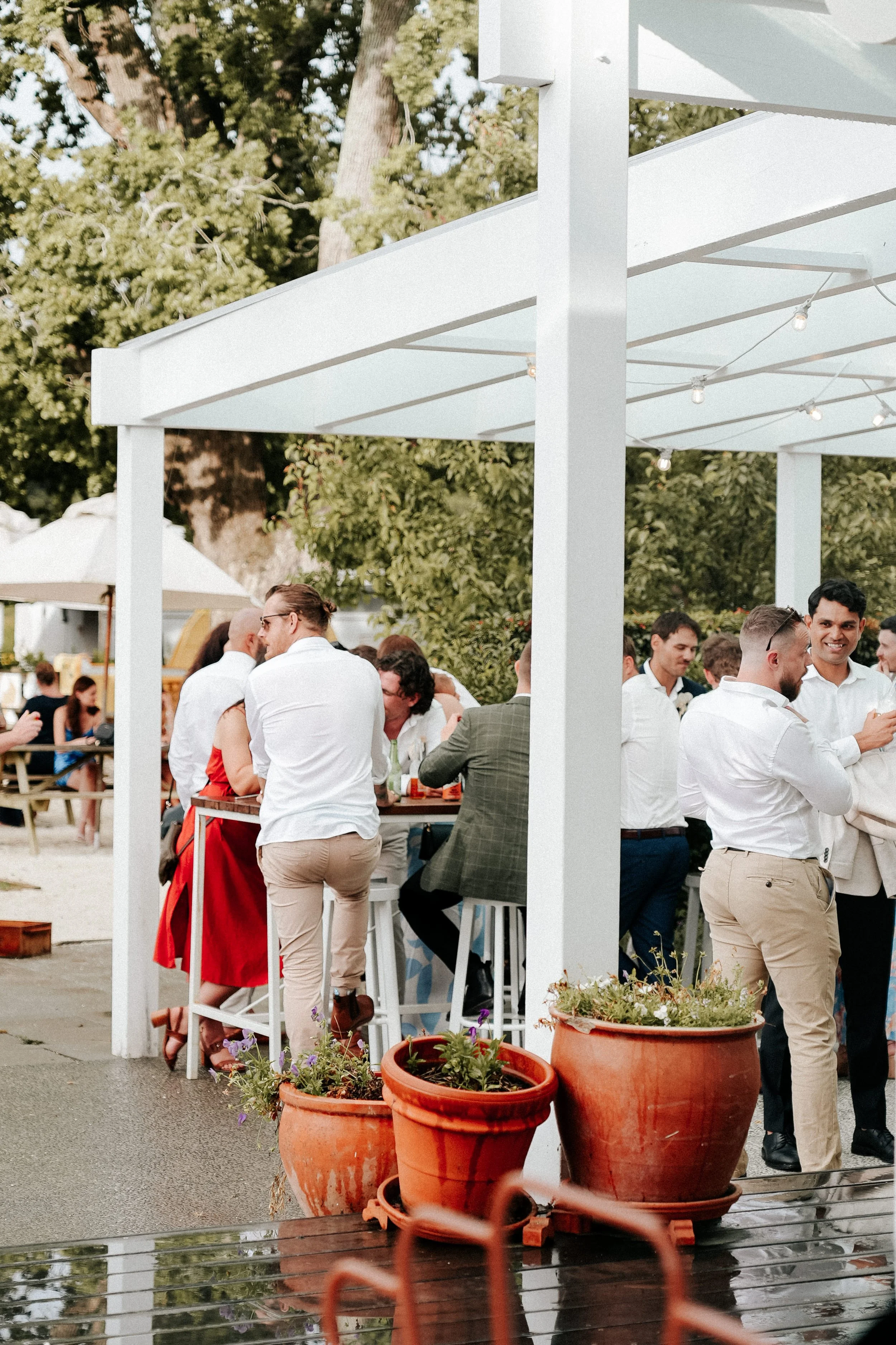 People socializing and chatting at an outdoor party under a white pergola with potted plants in the foreground.