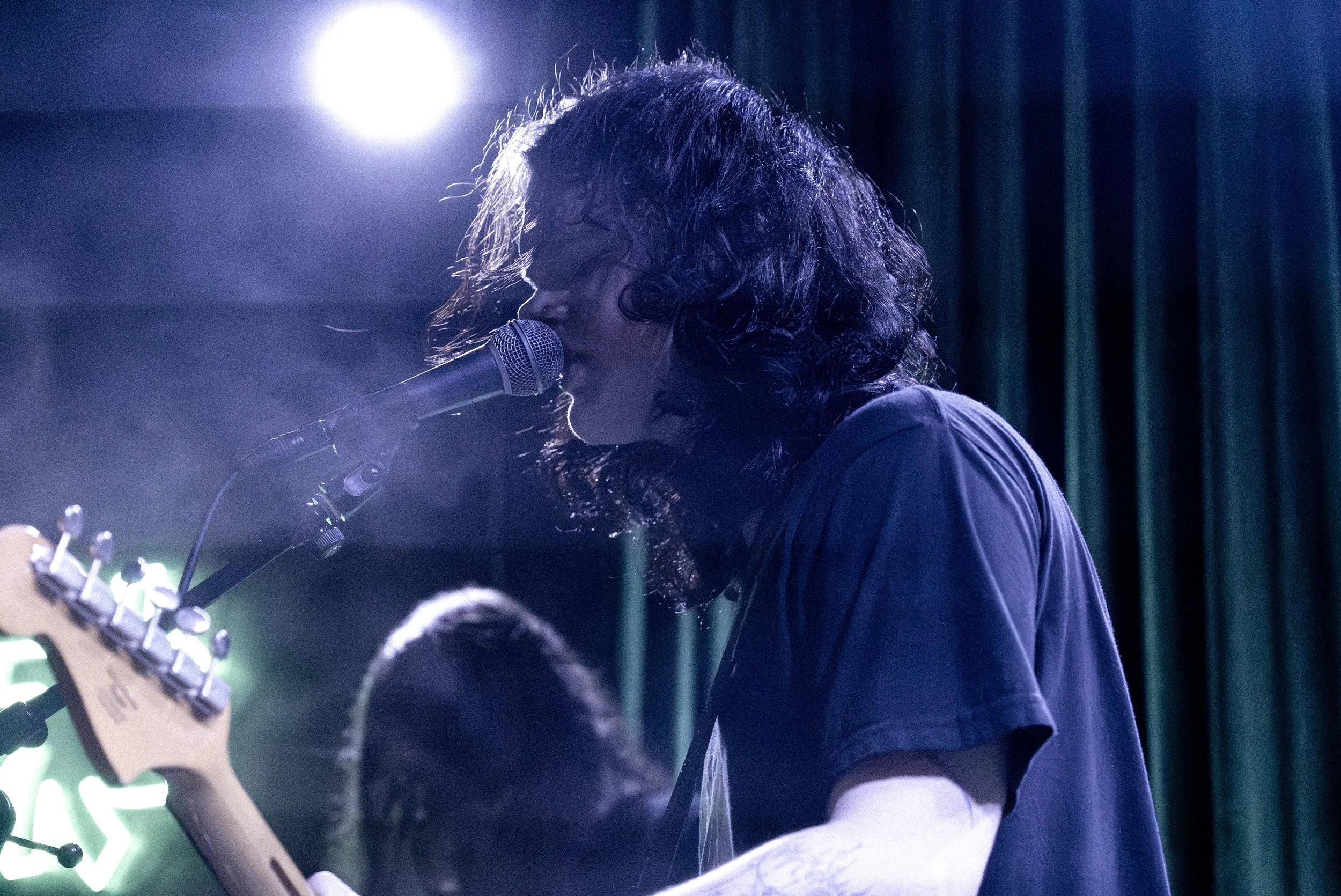 A musician with long, curly hair singing into a microphone while playing an electric guitar on stage under bright lights, with a dark backdrop.