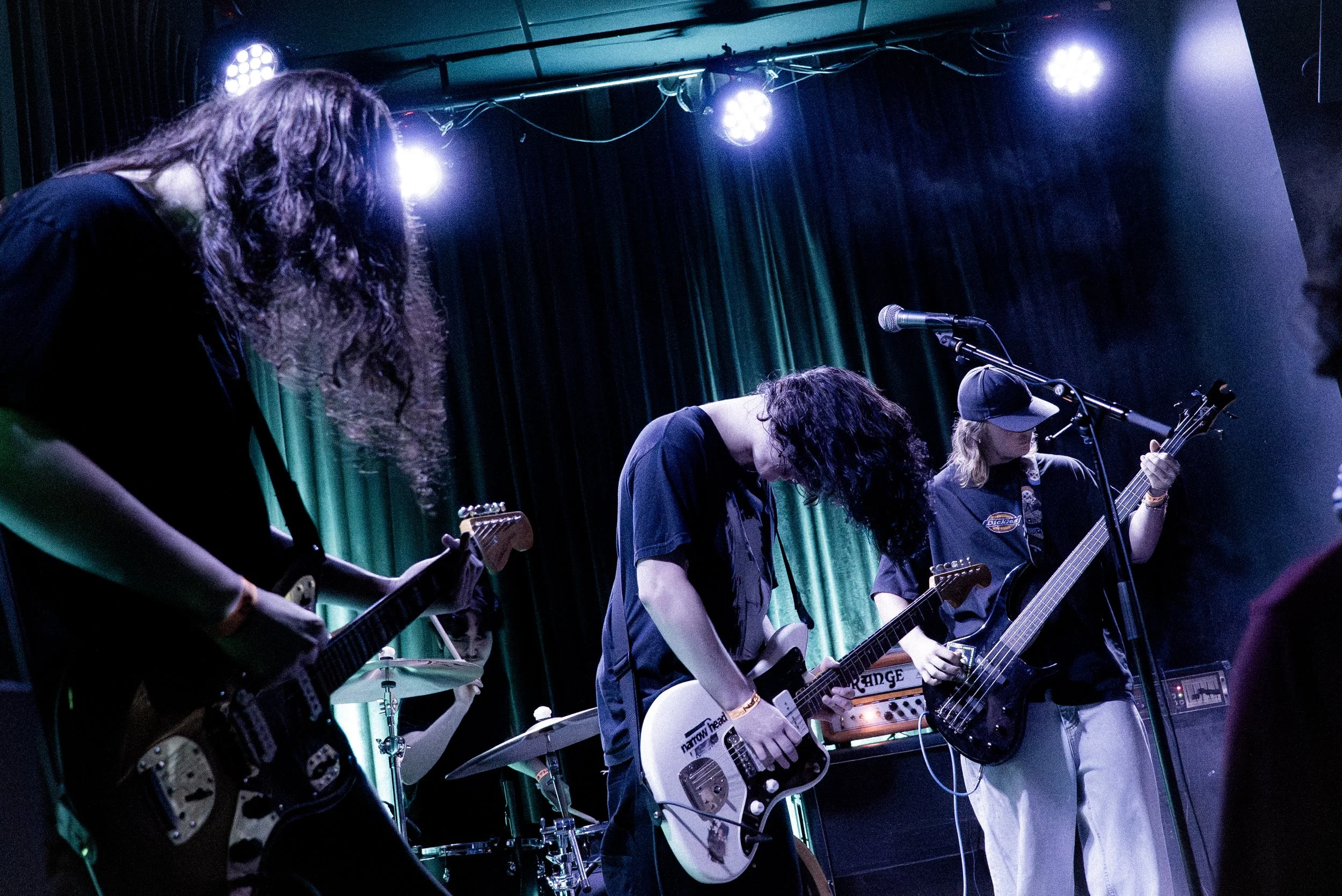A band performing on stage with three guitarists and a drummer, with blue lighting and green curtains in the background.