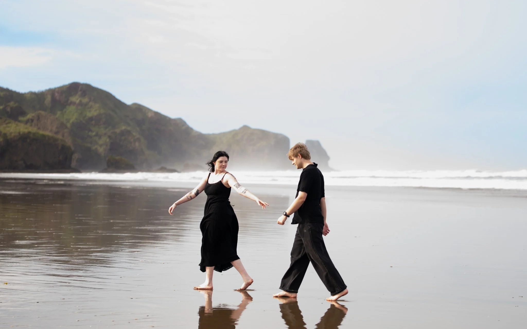 Two people walking barefoot on a wet beach with waves crashing behind them, green cliffs in the distance, and a partly cloudy sky.