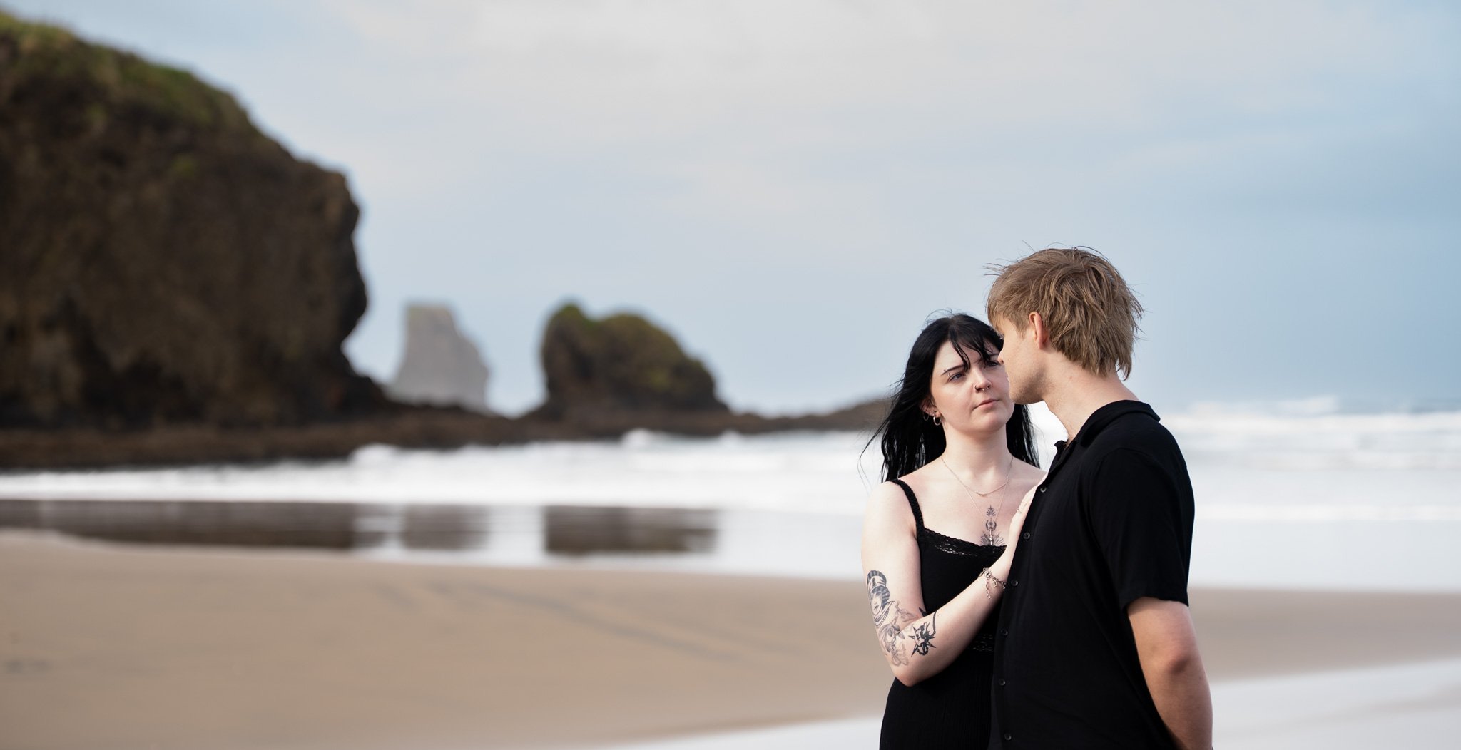 A couple standing close on a beach with rocky cliffs in the background, facing each other.