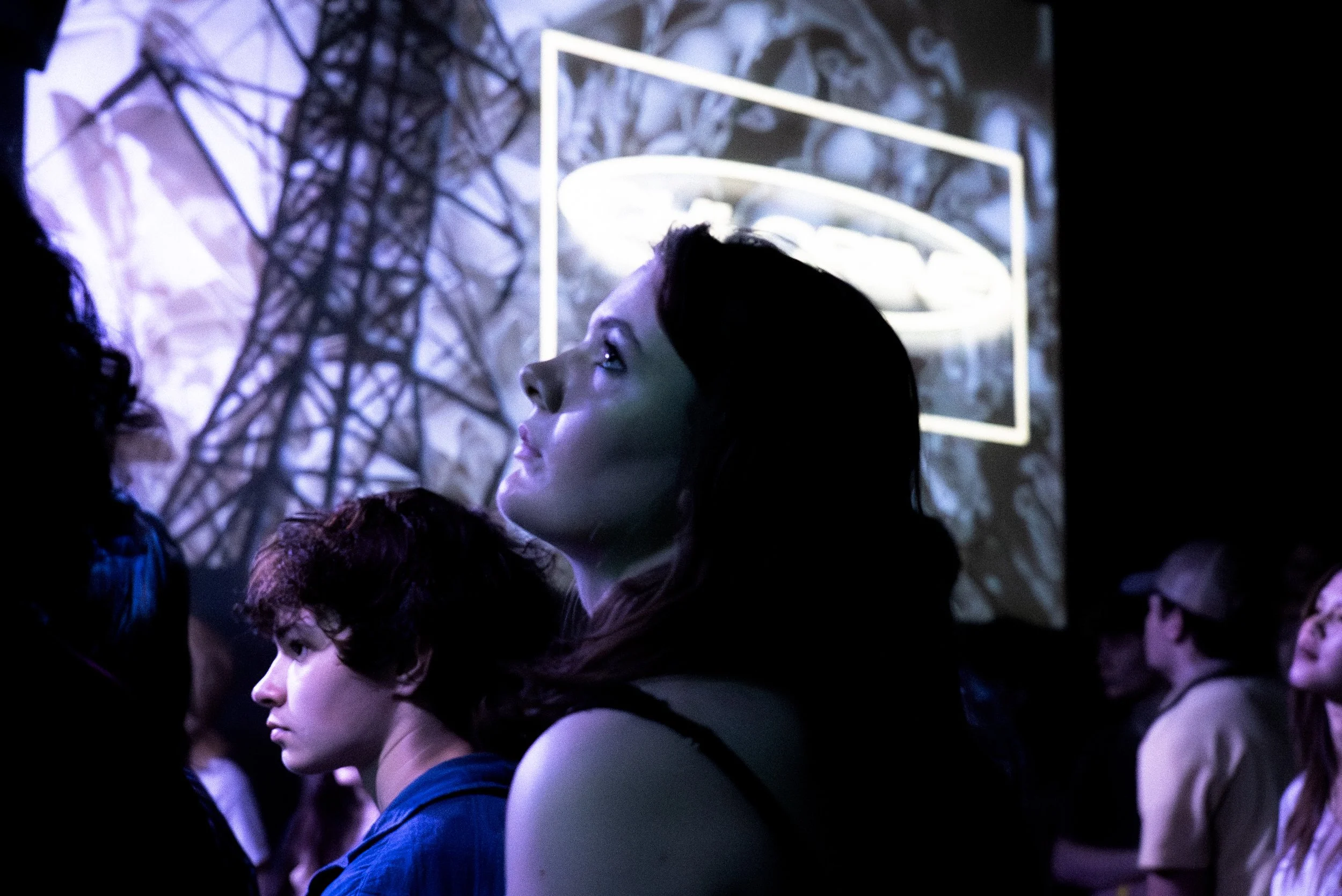 A group of young people attending a nighttime event, with a large screen showing an industrial structure in the background.