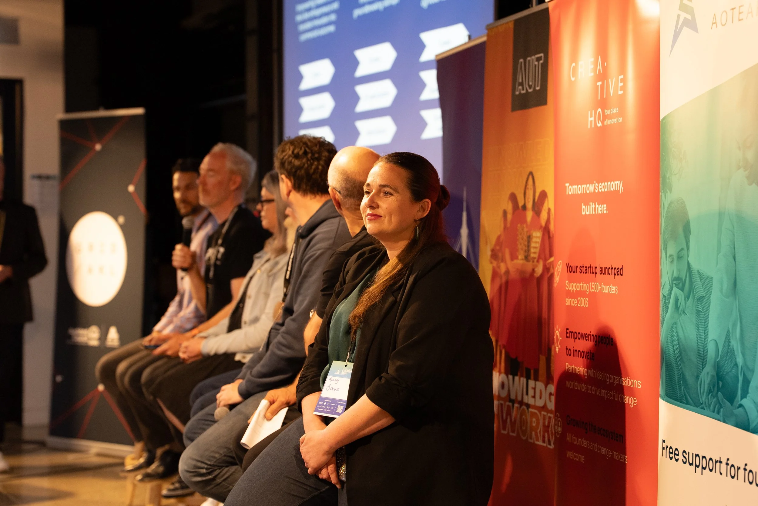 People sitting on a stage during a conference or panel discussion, with large colorful banners displaying event information behind them.