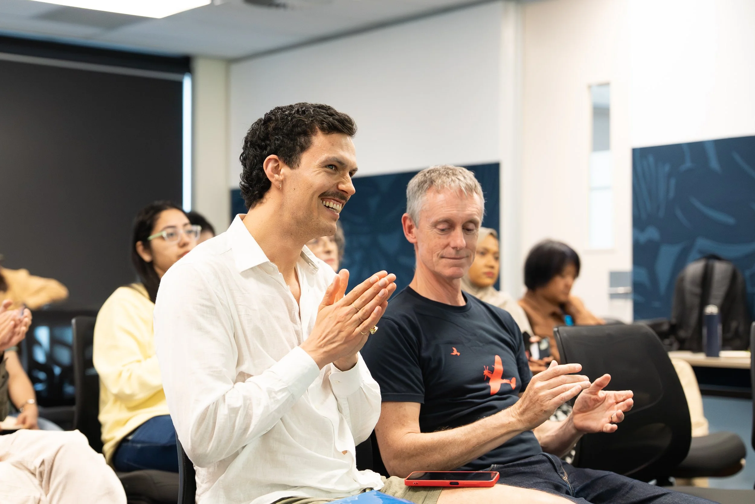 People attending a presentation or meeting in a conference room, with two men in the foreground clapping and smiling.