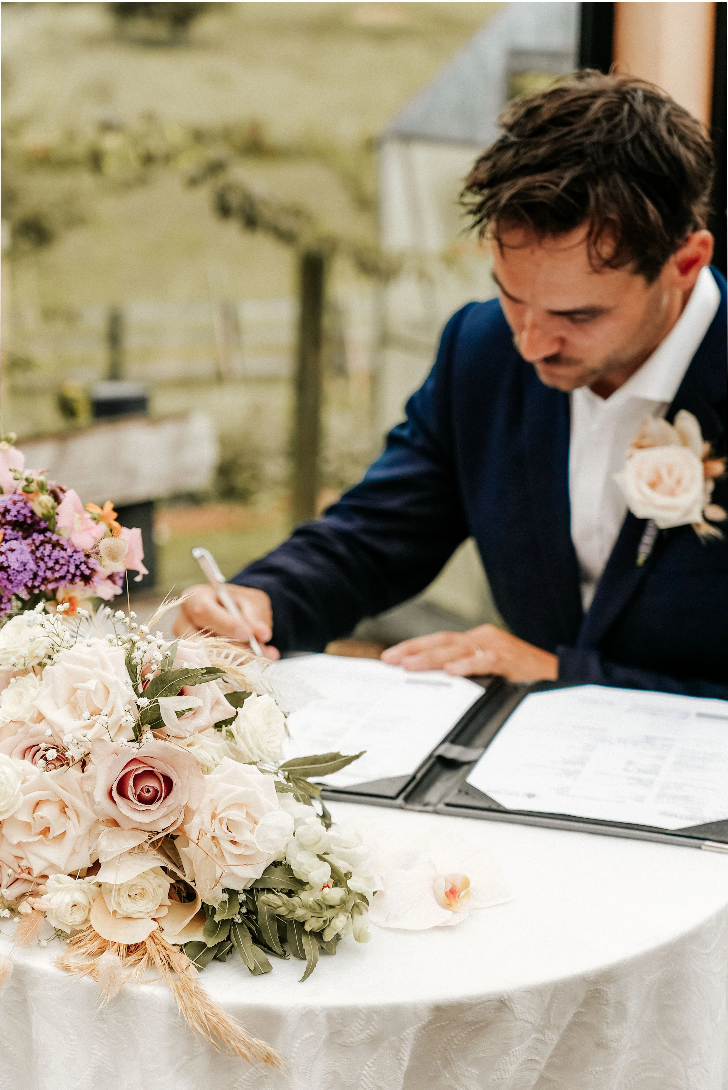 A groom in a navy suit signs a marriage certificate at a wedding reception. There is a floral bouquet with roses and other flowers on the table.
