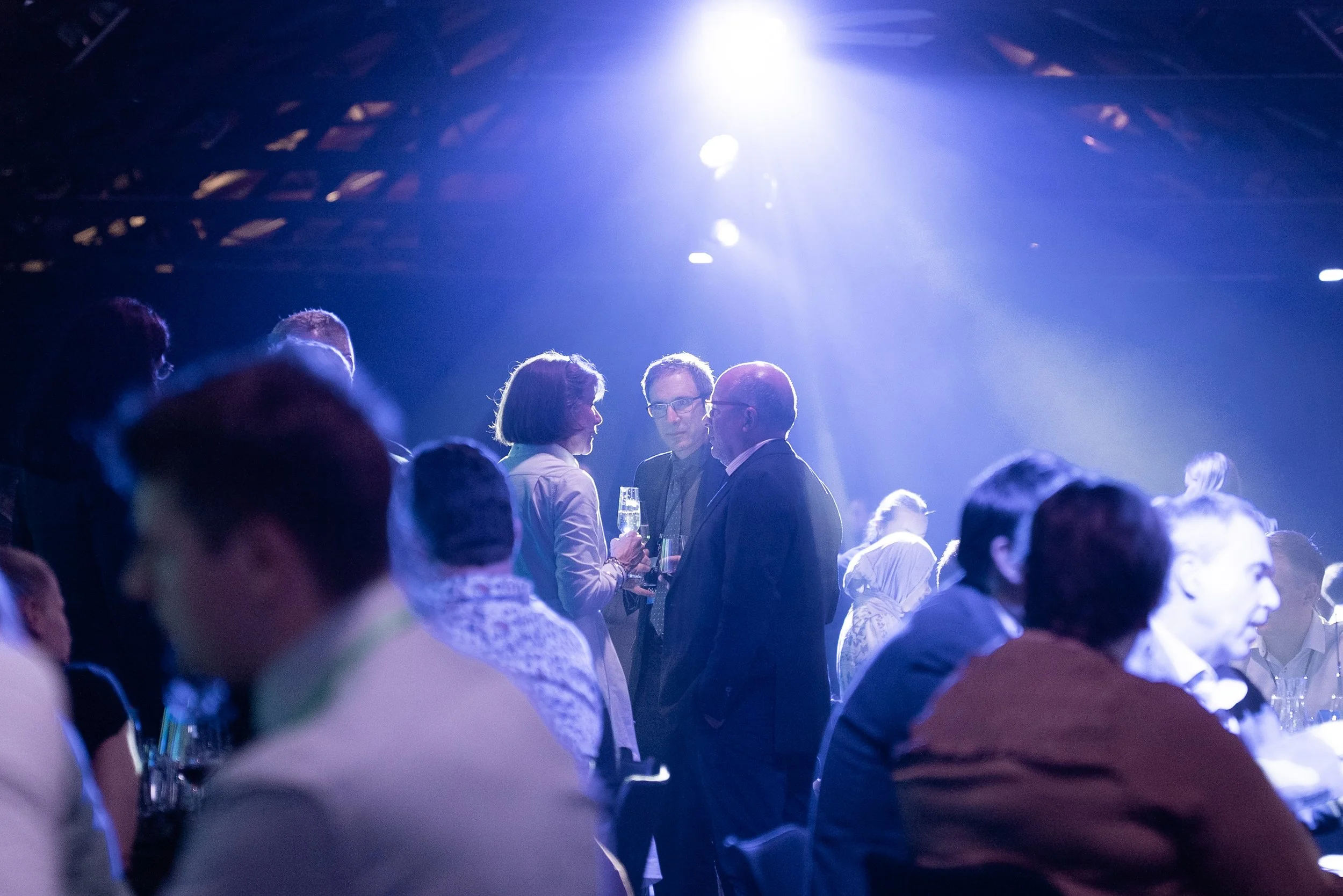 People socializing at a darkened event with blue lighting, engaged in conversation, some holding glasses of champagne.