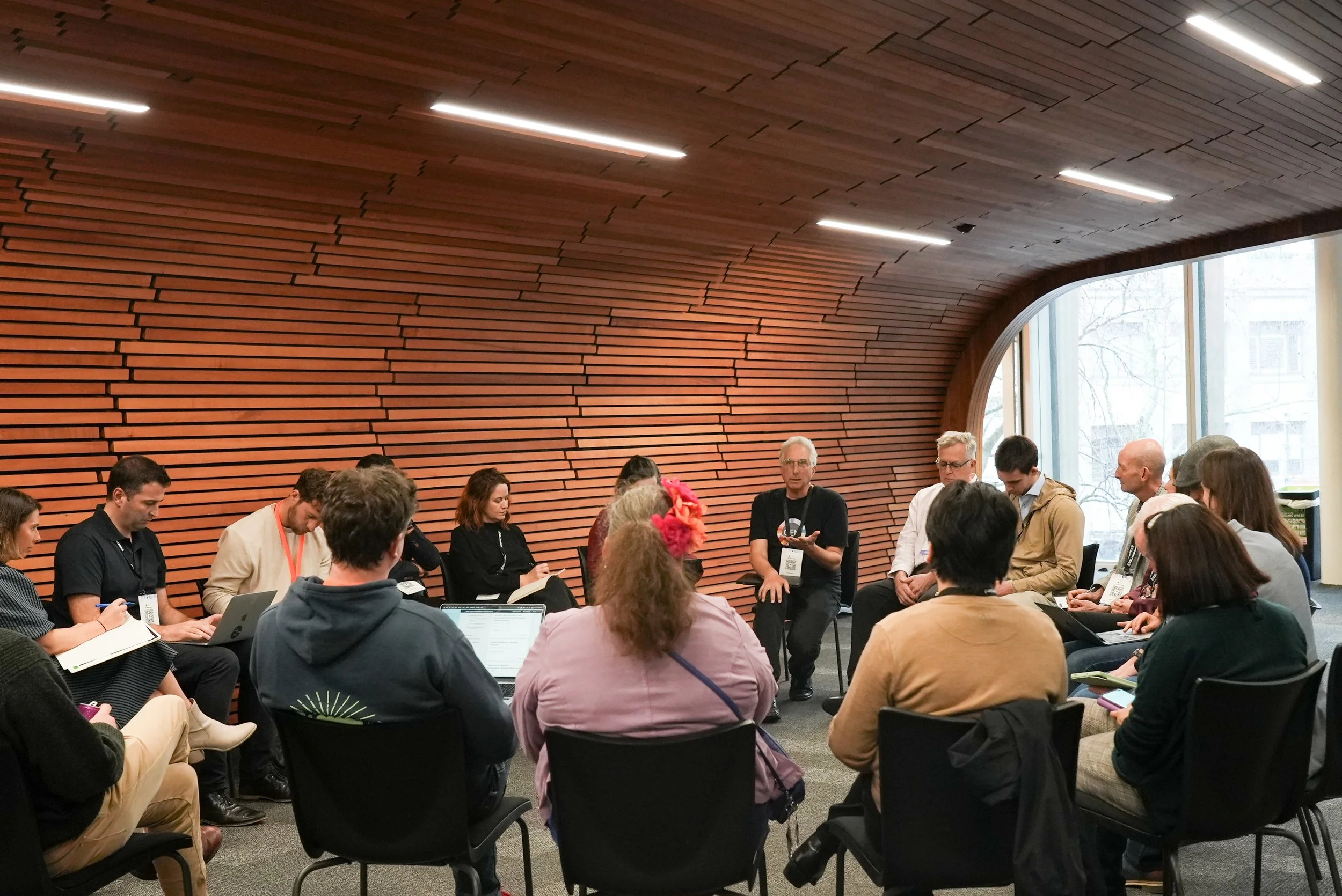 A group of people sitting in a circle in a modern room with wooden wall panels and large windows, participating in a discussion or workshop.