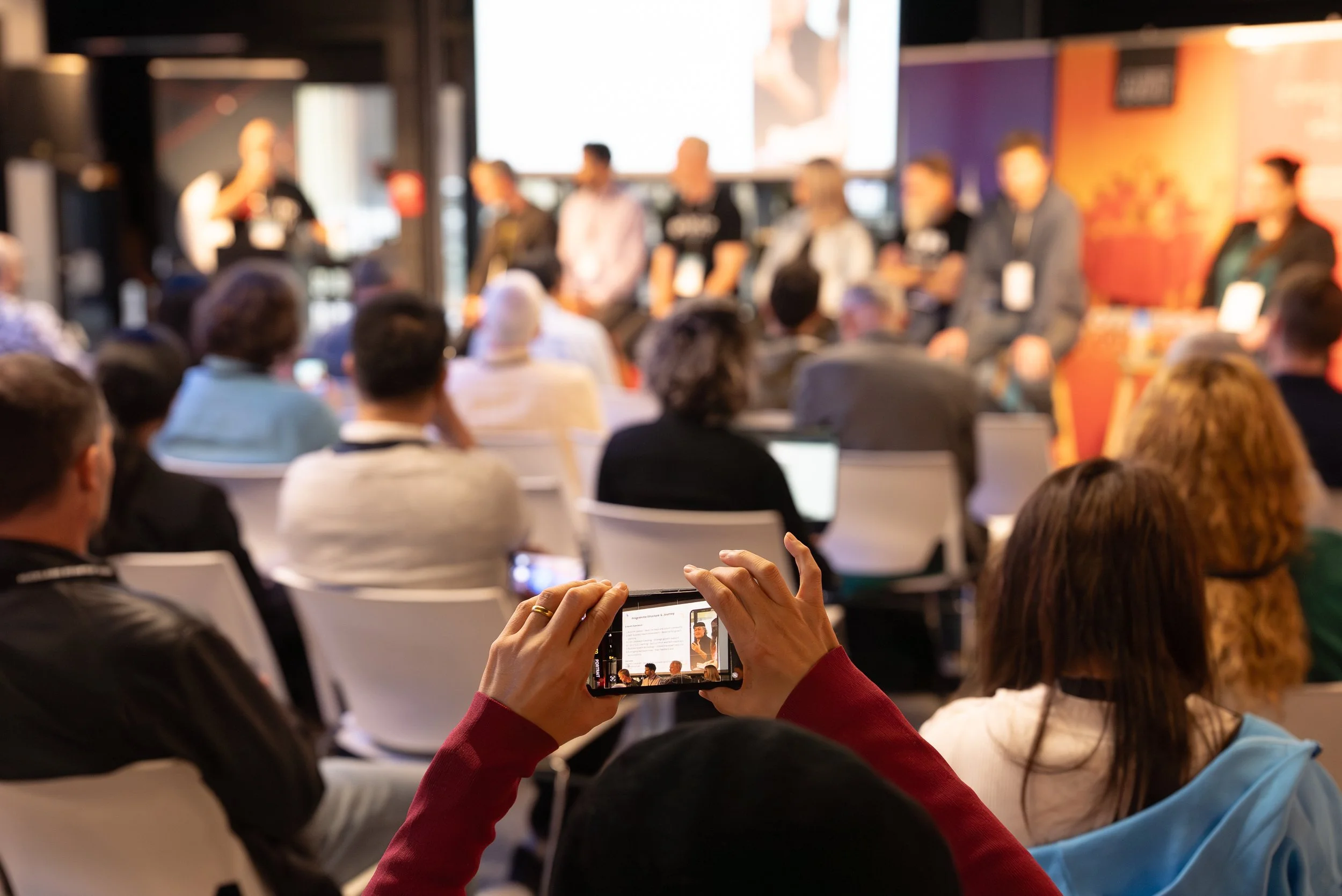 A person in a red shirt is taking a photo of a panel discussion at a conference with their smartphone. The audience is seated, and the panelists are sitting on stage in front of a large screen.