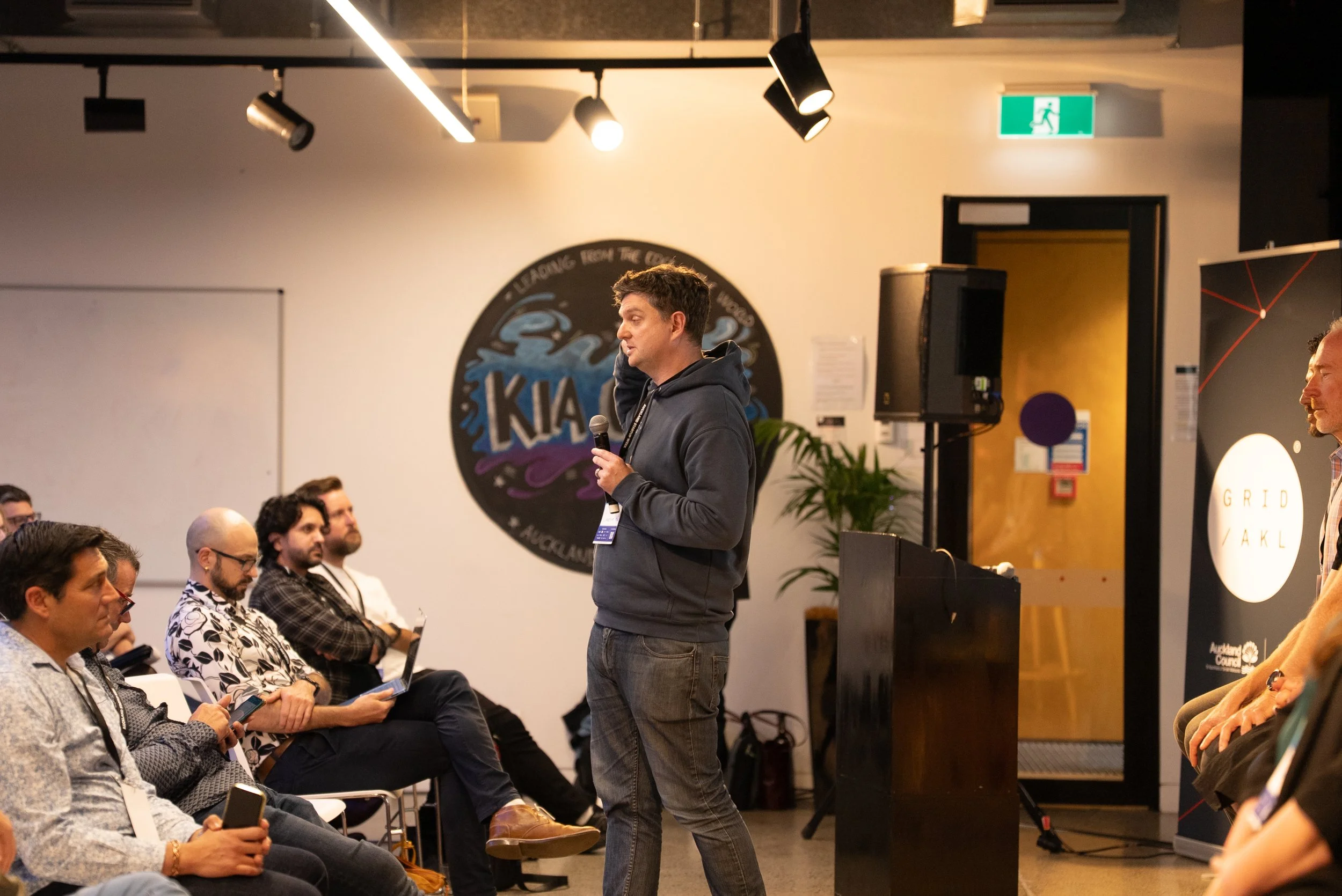 A man in a dark hoodie is giving a presentation with a microphone in a conference room. Several attendees sit and listen, some using laptops or smartphones. The room has a logo on the wall behind him that reads 'KIA' with a wave design, and there is a speaker and a plant near the door.
