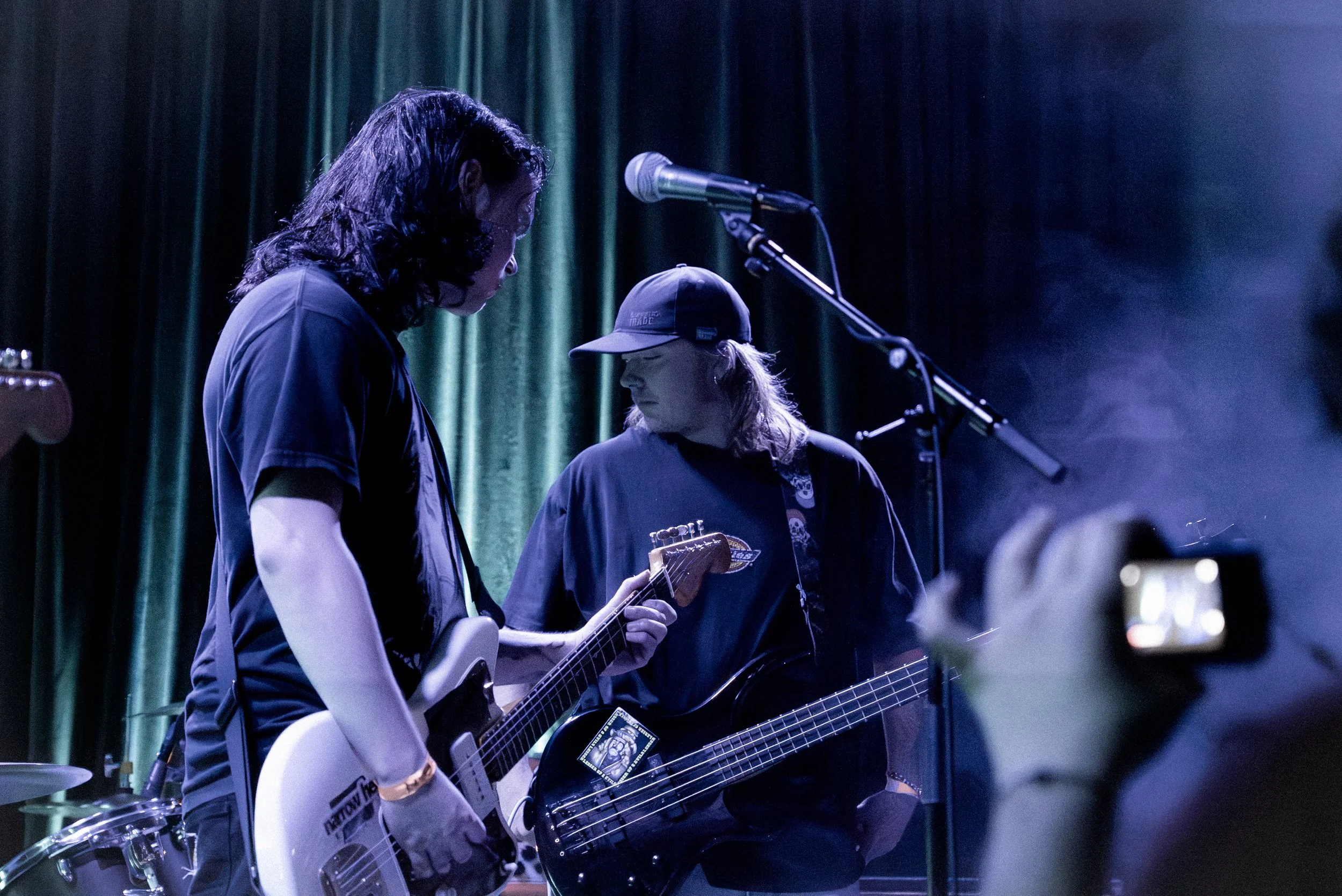 Two musicians with long hair and casual clothing performing on stage, with a green curtain backdrop and a microphone stand nearby.