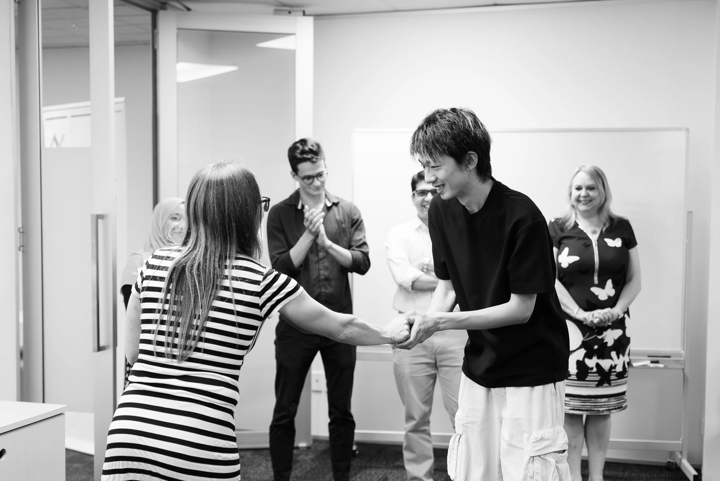 A young man and woman shake hands in a congratulatory gesture while others look on and smile in a room with white walls and a whiteboard.