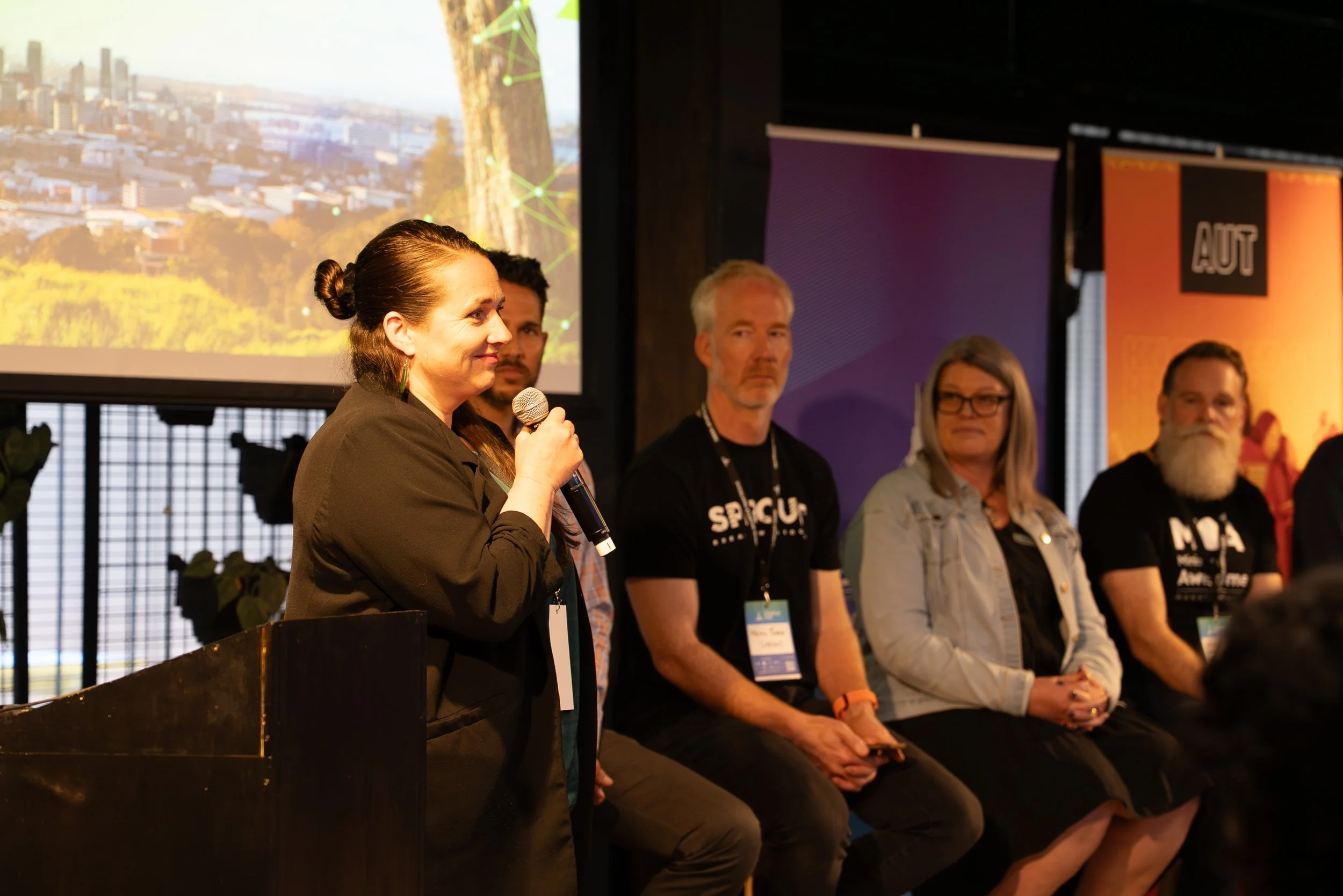 Speaker at a panel discussion holding a microphone, seated next to four other panelists, with a large screen displaying a cityscape behind them at a conference.