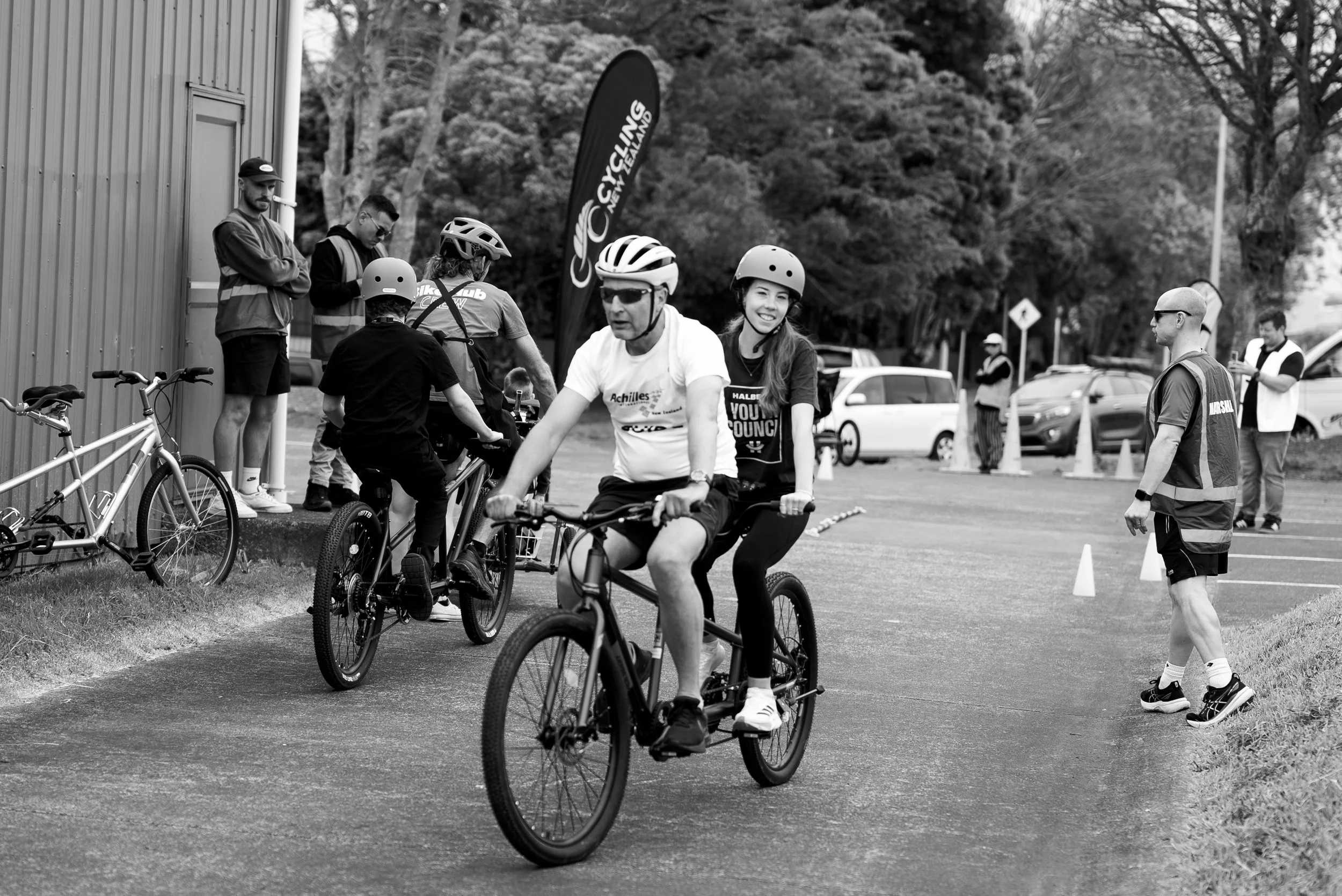 People riding bicycles at an outdoor cycling event, with spectators and event staff watching.