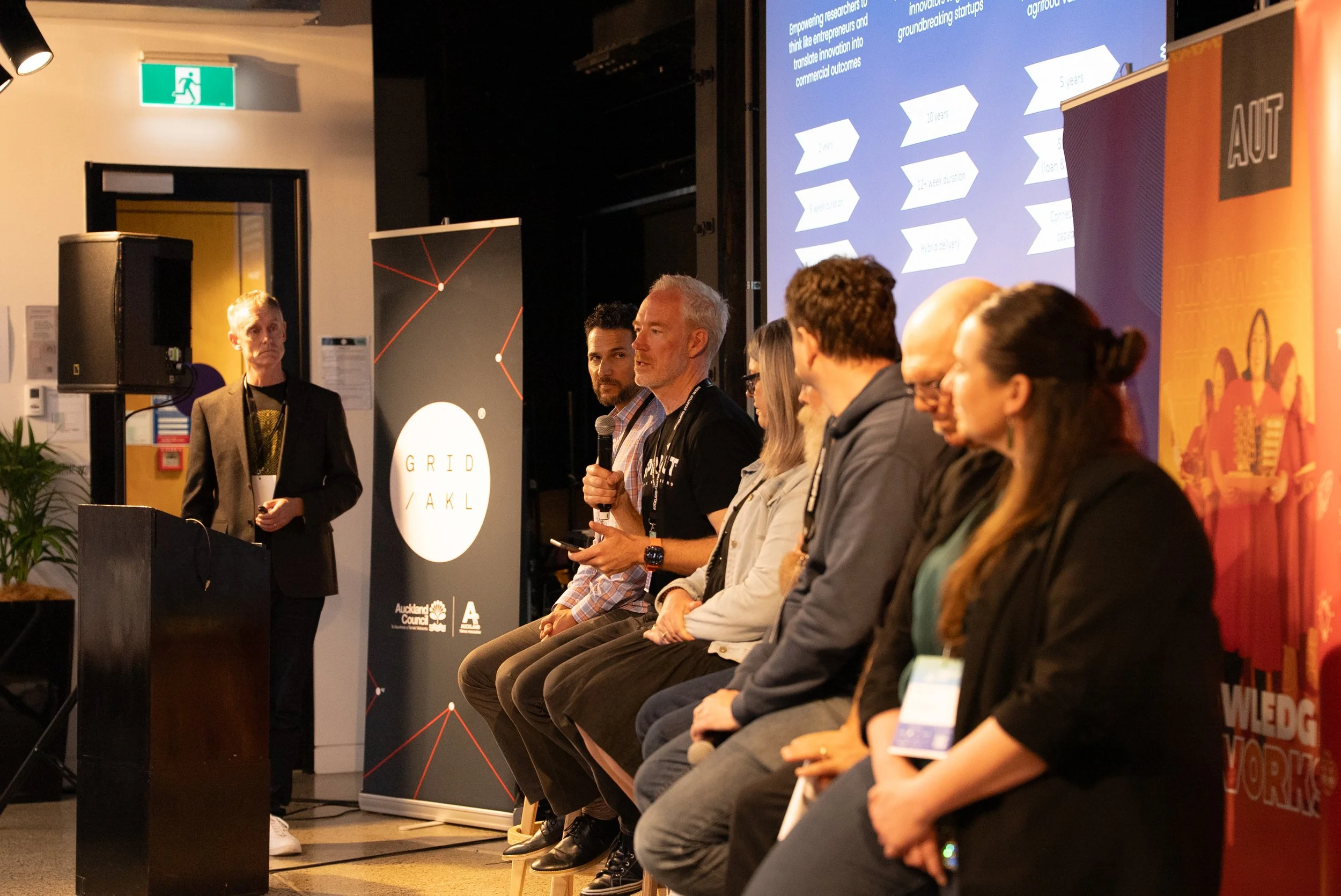 A panel of seven people sitting on chairs at a conference, with one person standing near a podium. One of the panelists is speaking into a microphone. Behind them are banners and a large digital screen displaying text and graphics. An emergency exit sign is visible above a door in the background.