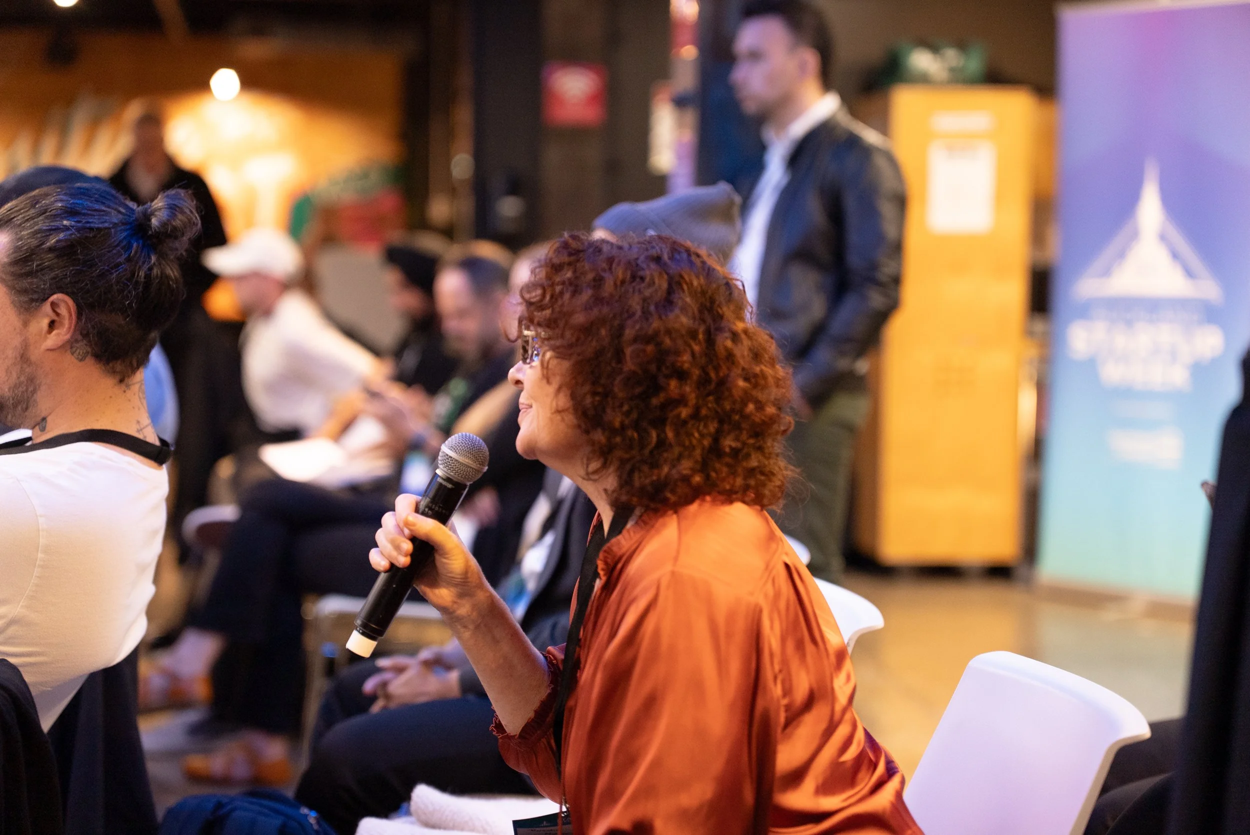 A woman with curly red hair and glasses, wearing an orange jacket, is holding a microphone and speaking at an indoor event. There are several people sitting in chairs around her, some of whom are looking at their phones. In the background, a man with short dark hair, wearing a leather jacket, is standing. A large blue sign with a white logo and text is partially visible to the right.
