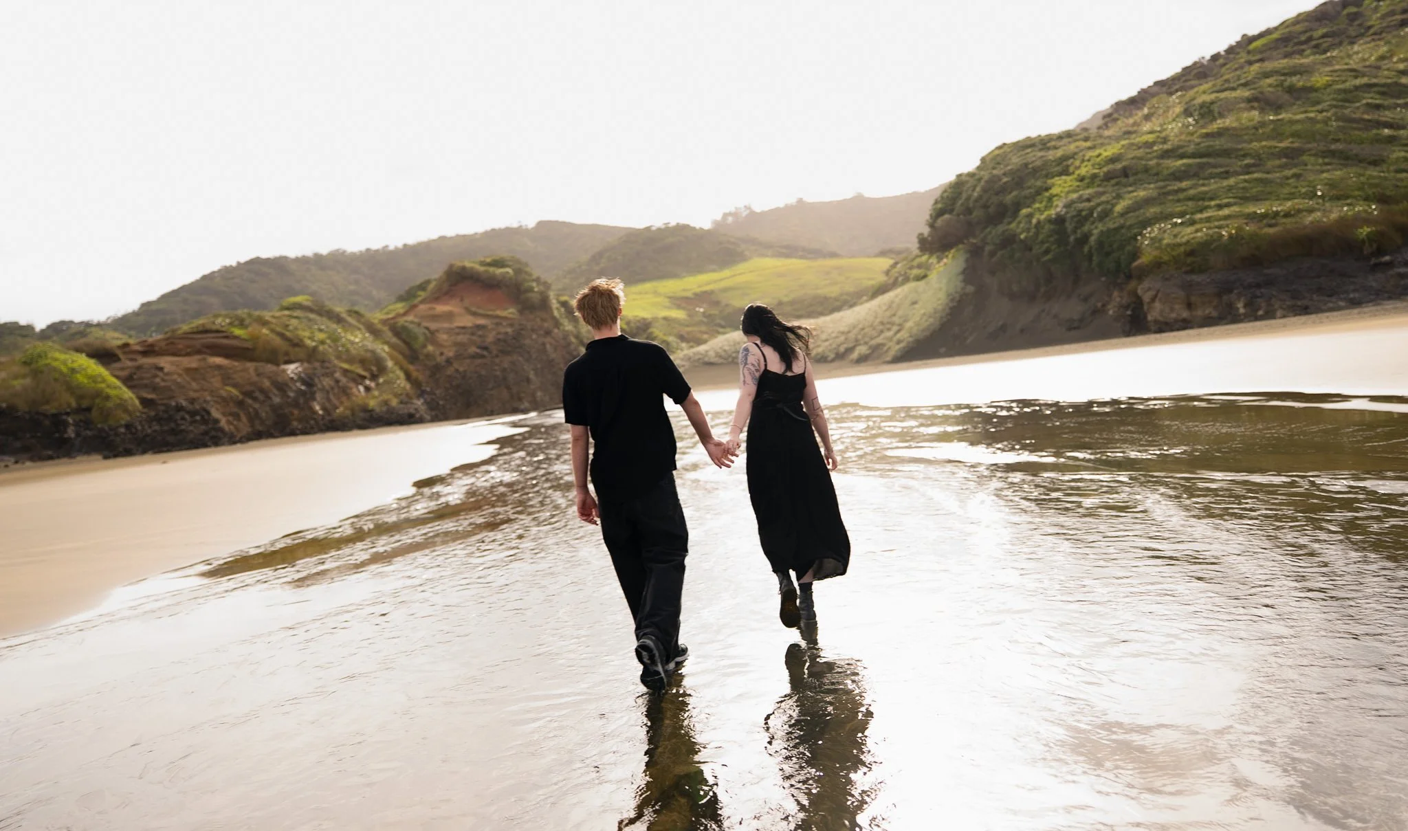 A couple holding hands walks on the wet sand of a beach with green hills in the background.