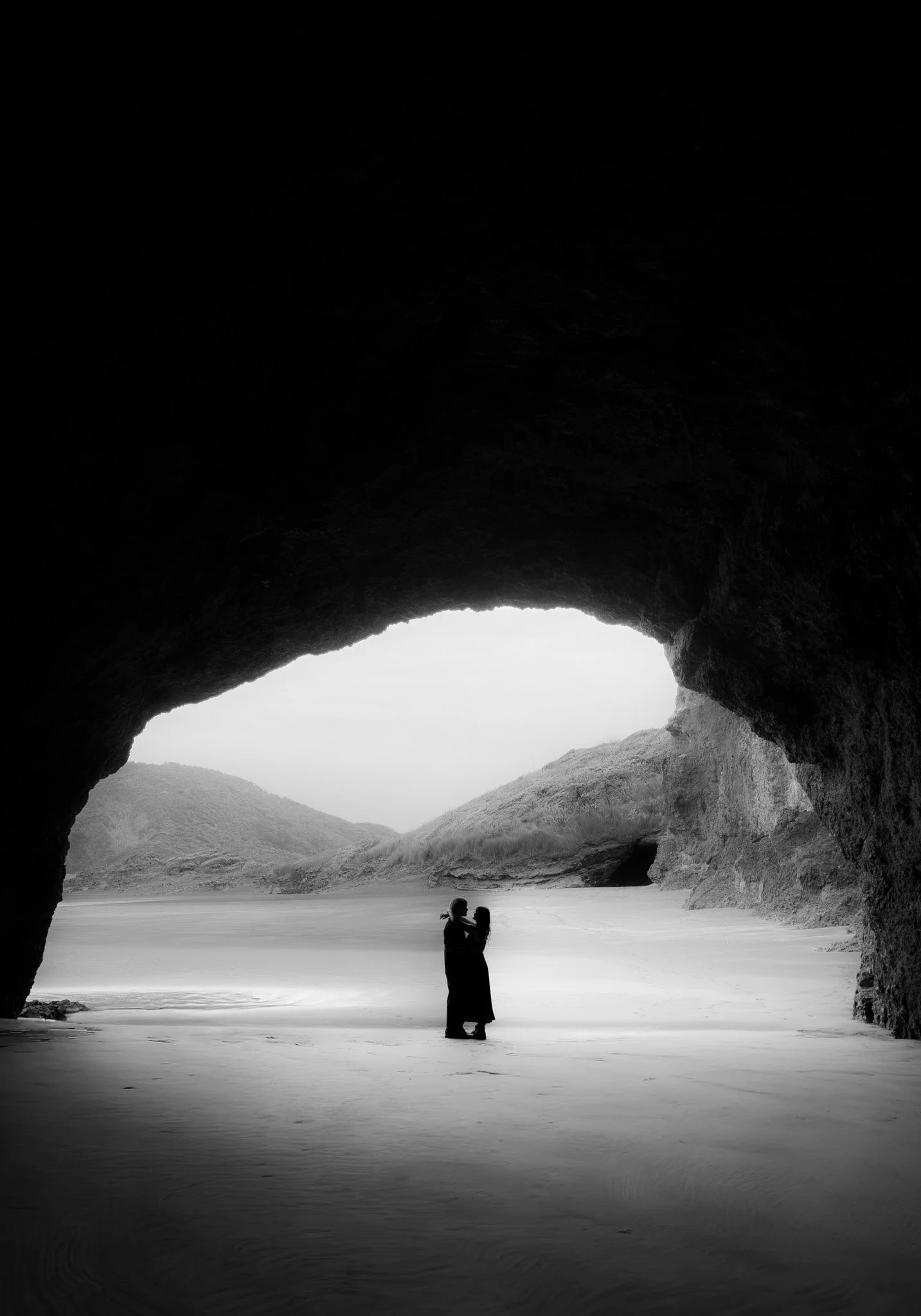 A black-and-white photograph of two people standing close together in a cave opening, with a landscape of hills and water in the background.