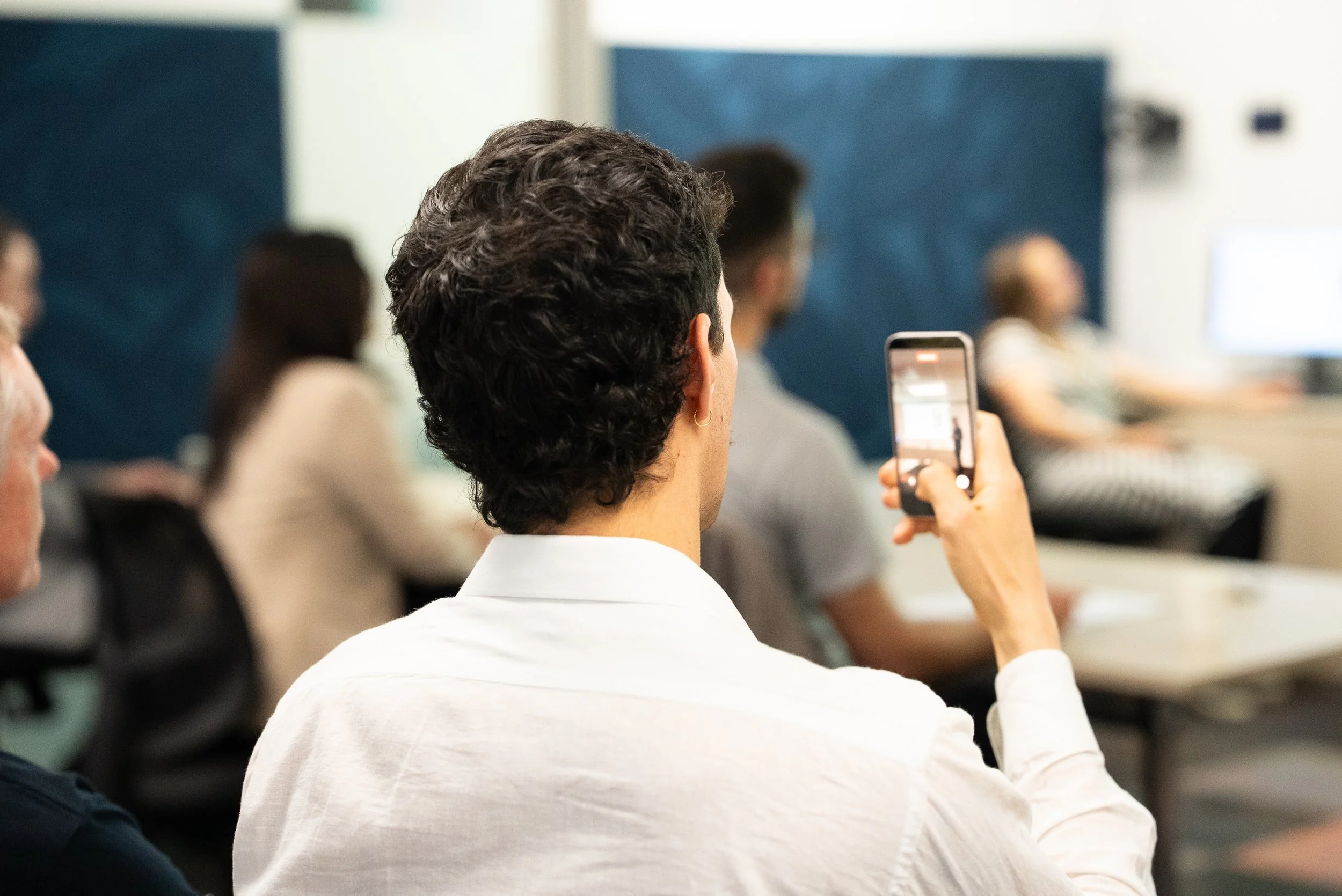 A person with short, curly hair taking a photo or video with a smartphone during a seminar or conference in a classroom setting.