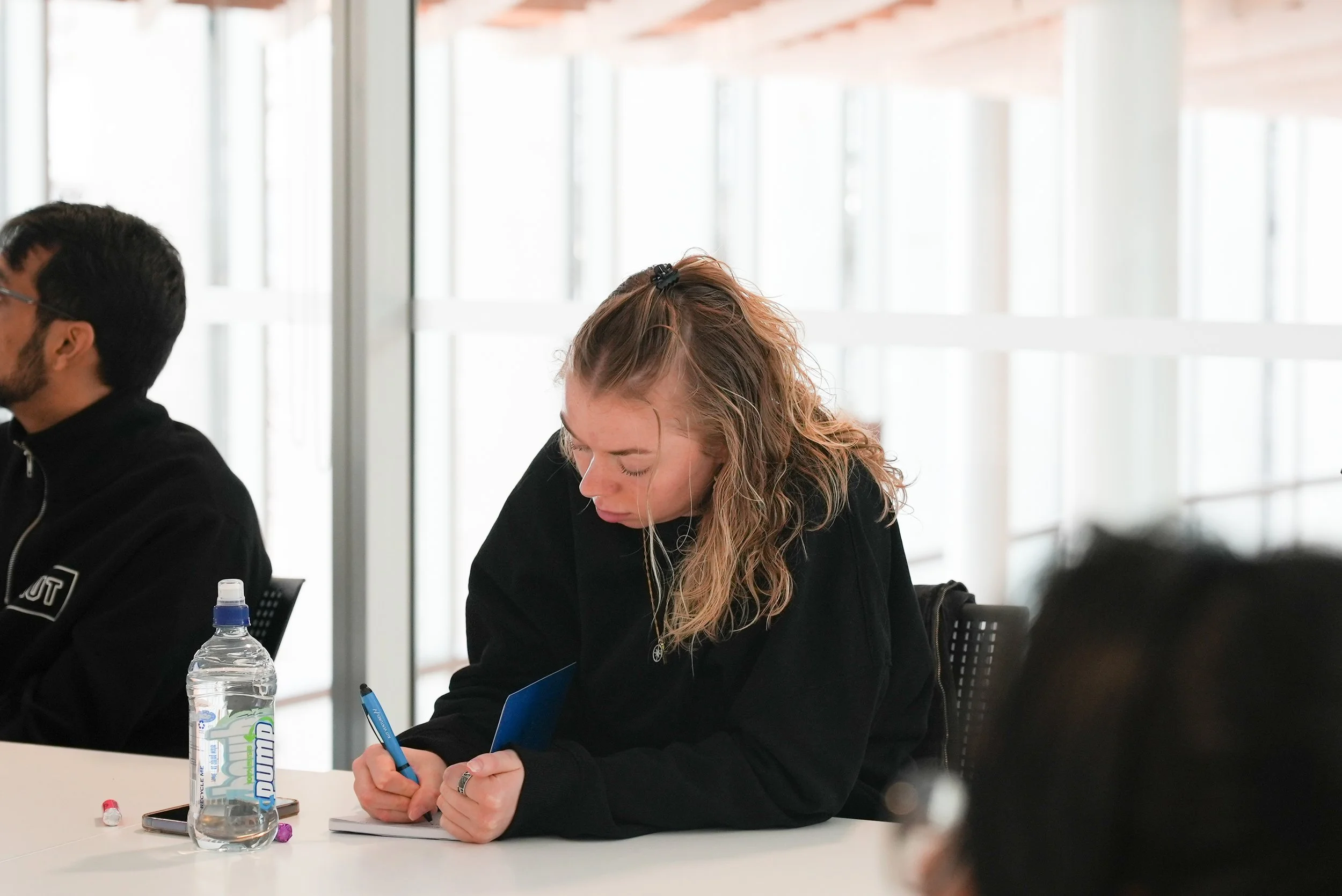 Young woman with curly blonde hair writing on a notepad in a conference room with large windows.