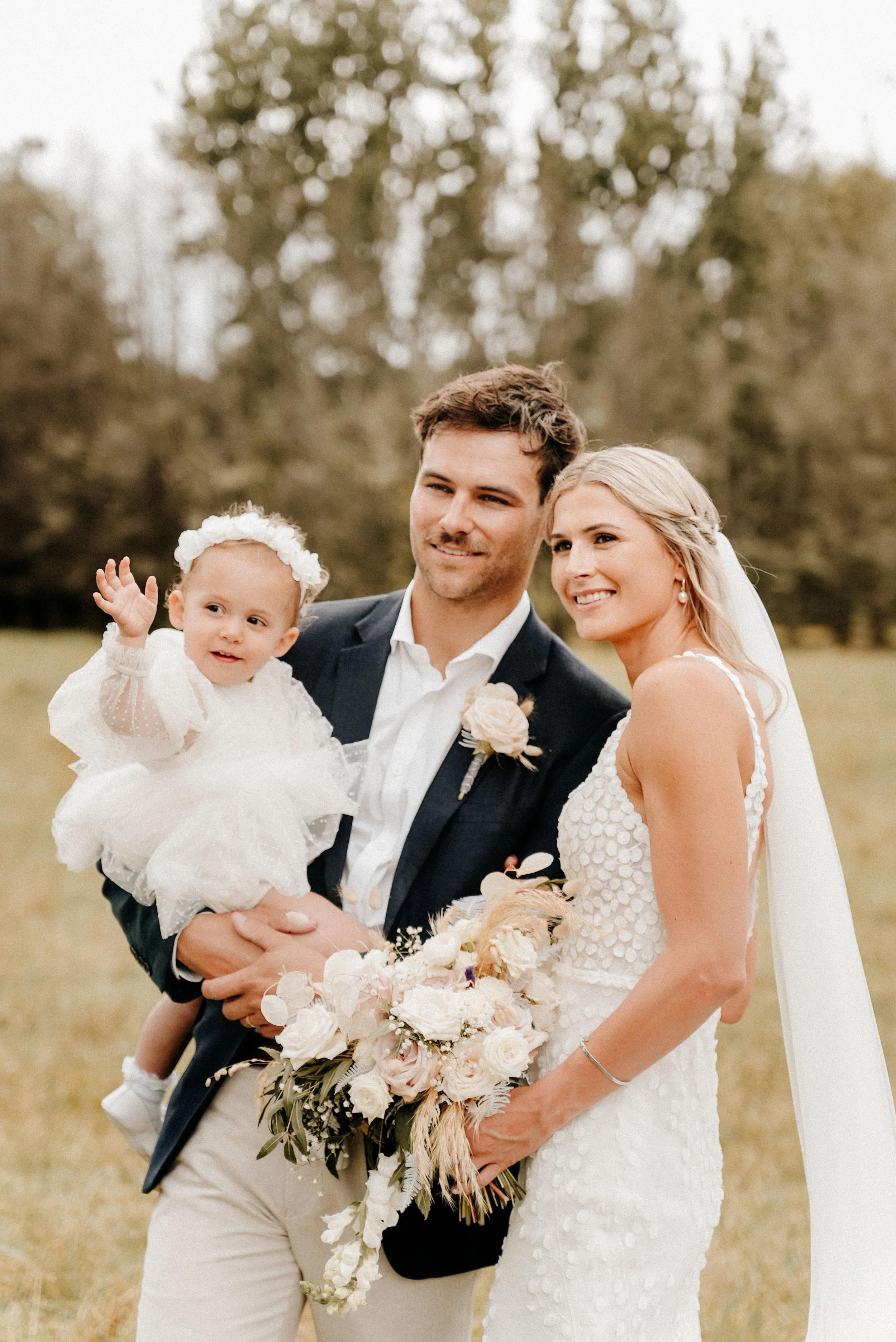 A wedding couple with a young girl outdoors during daytime. The bride is wearing a white wedding dress, holding a bouquet of flowers, and has long blonde hair. The groom is dressed in a navy jacket and white shirt. The little girl, dressed in a white dress with a floral headband, is being held by the groom and is waving.