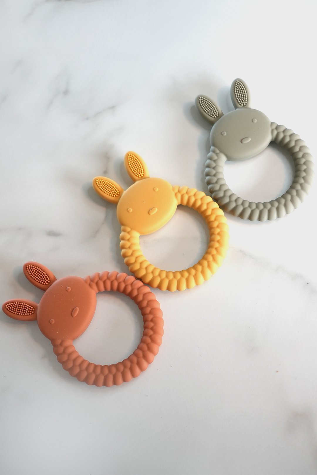 Three bunny-shaped silicone teething rings in pink, yellow, and gray arranged on a white marble surface.