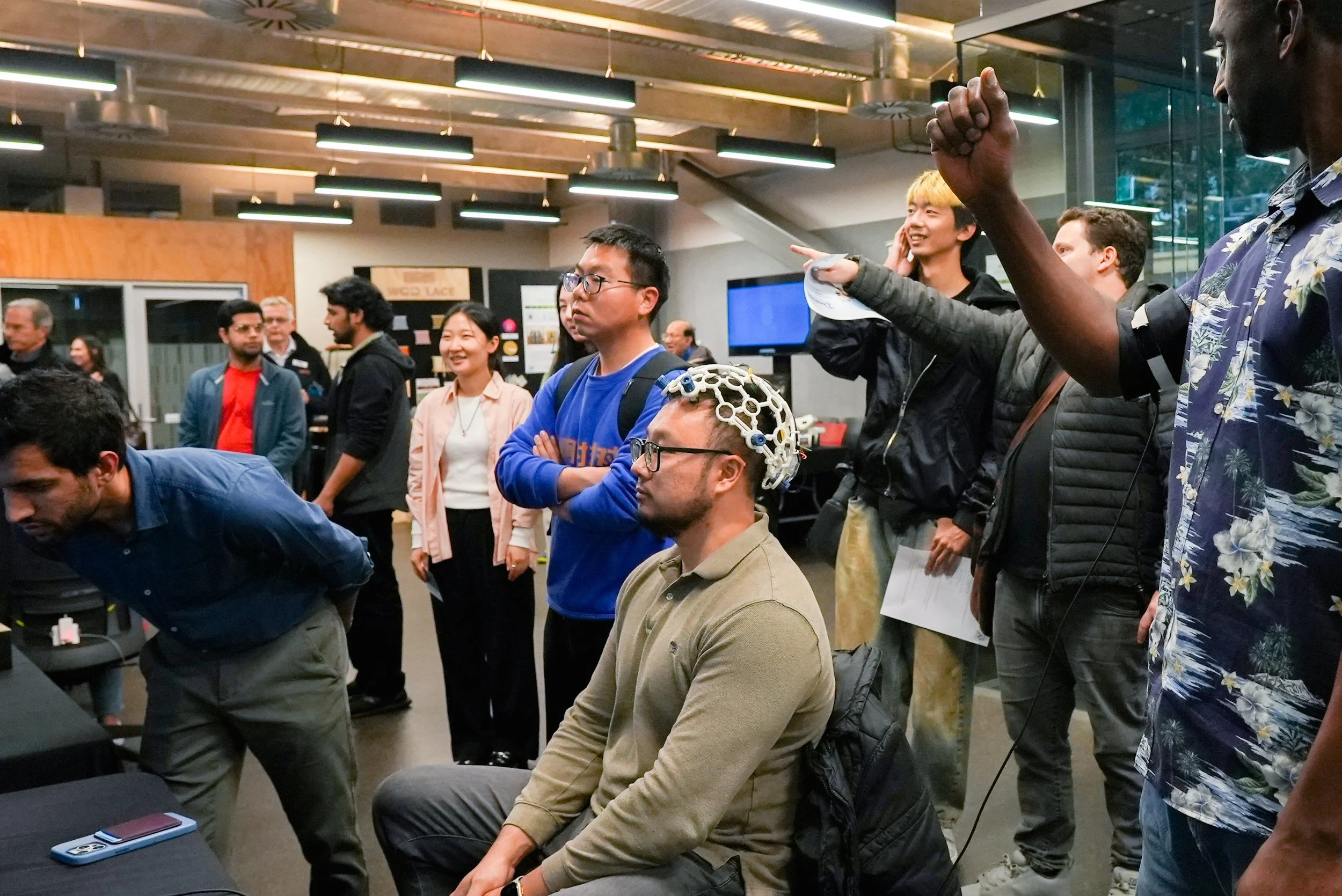 Group of people at a technology event, some wearing headsets, one person with a robotic headband, others standing and watching.