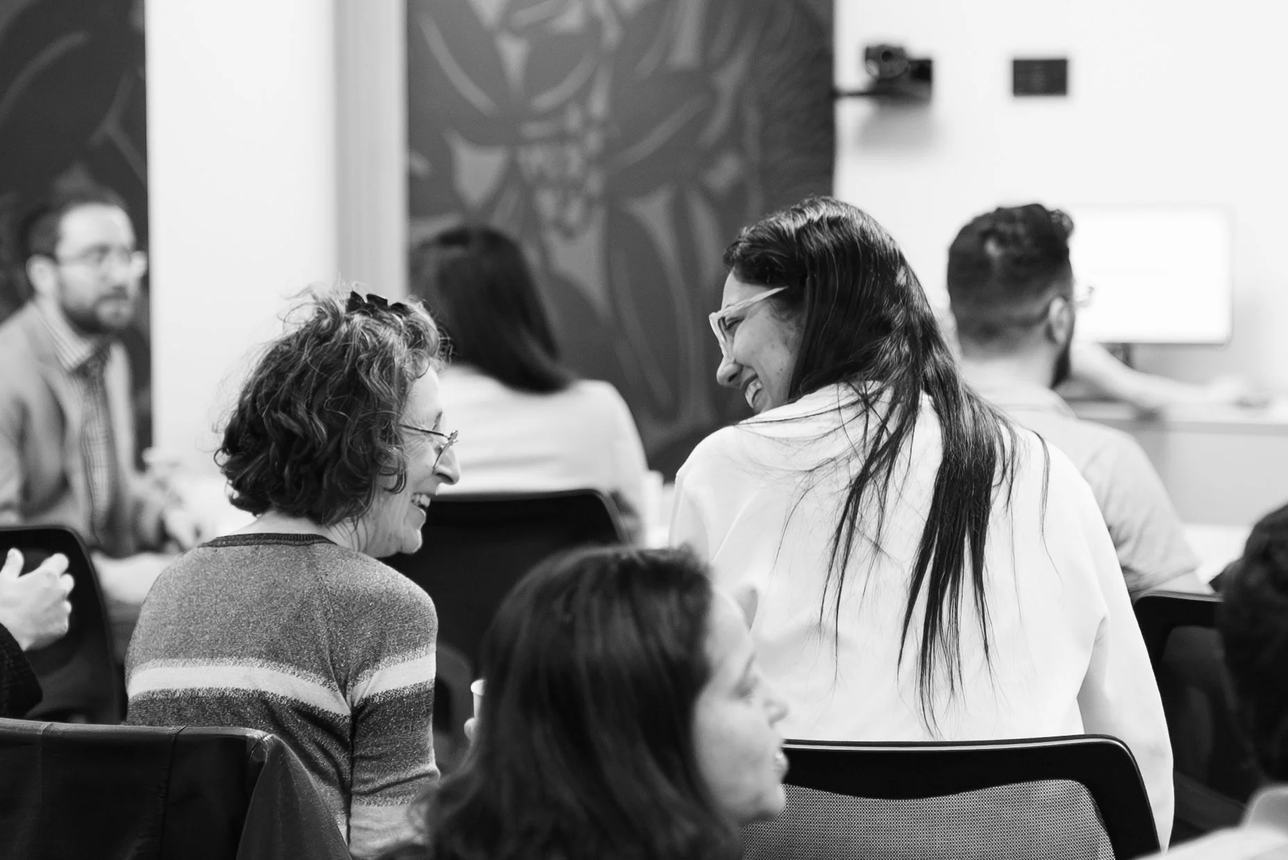 Two women in glasses are smiling and talking to each other in a conference room with several other people seated nearby.