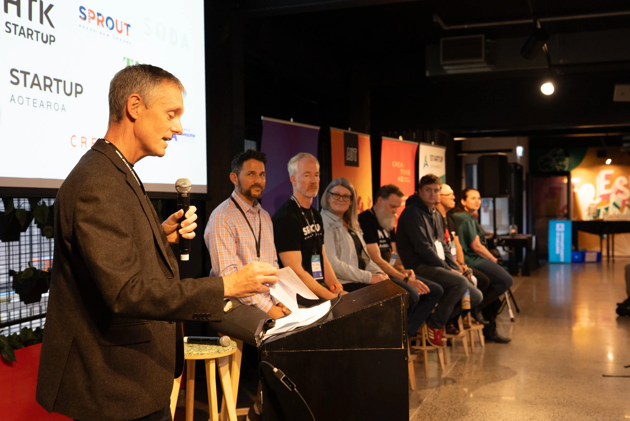A man in a brown blazer speaking into a microphone at a panel discussion, with six people sitting behind him on a stage, and a large screen displaying startup logos in the background.