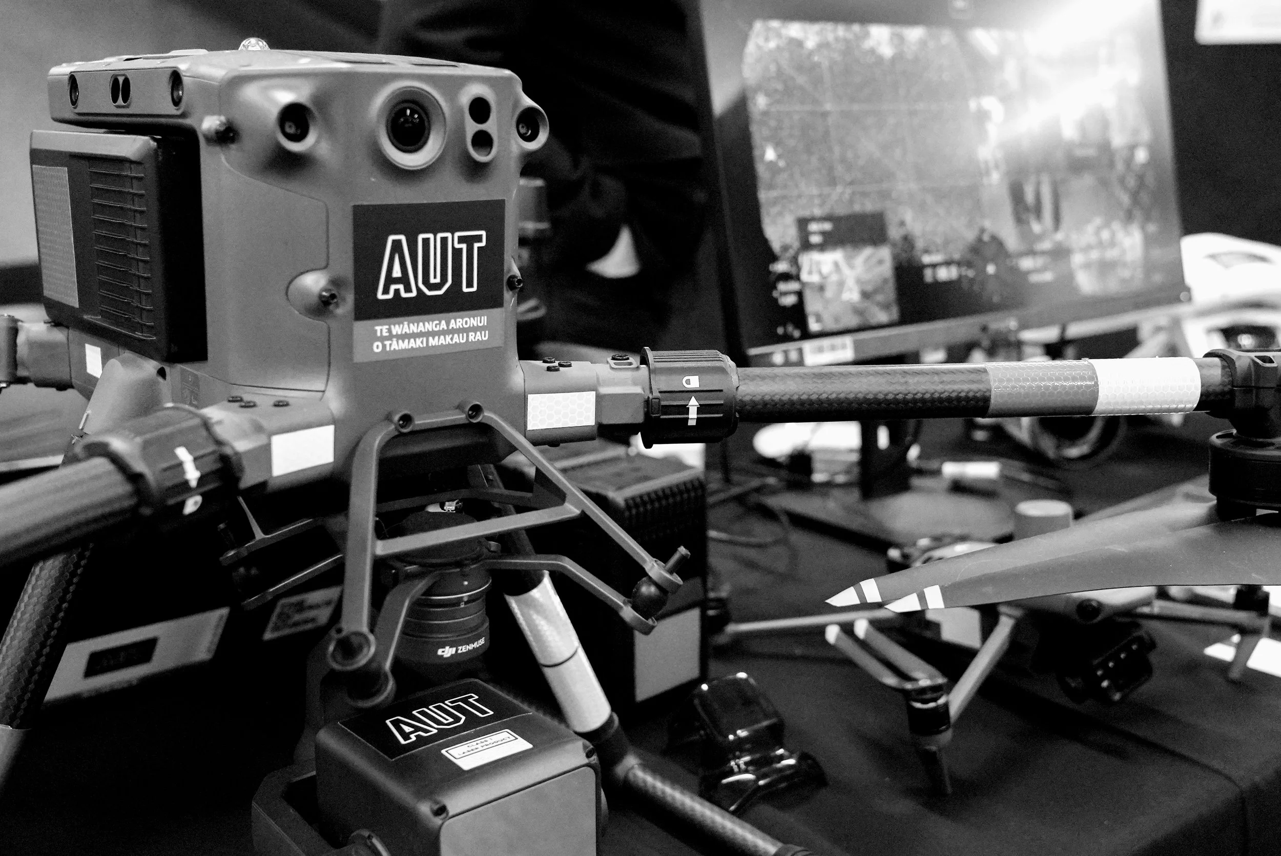 Close-up of a drone equipped with a camera and sensors on a desk, with a computer monitor in the background.
