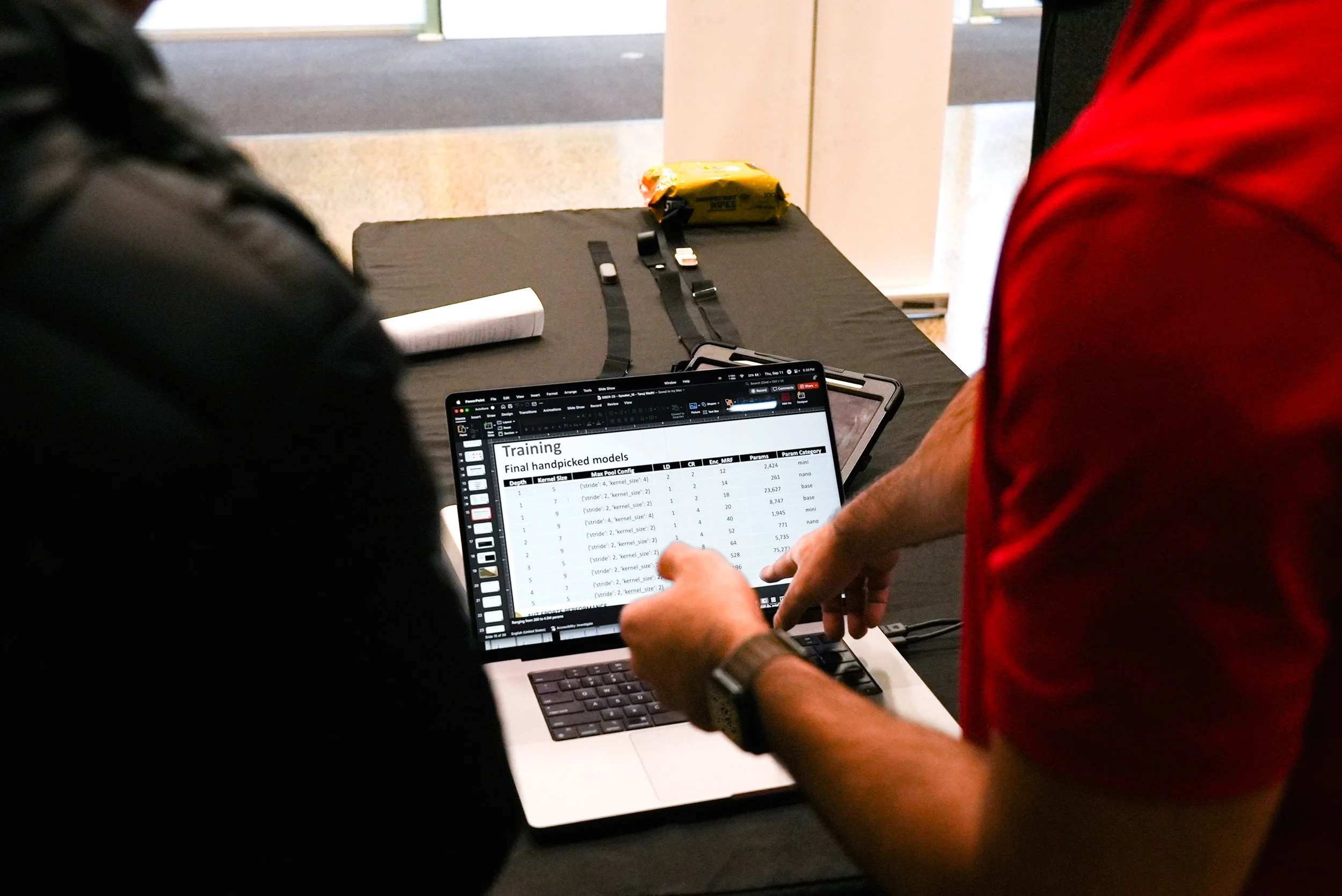 Two people looking at a laptop screen displaying a spreadsheet titled 'Training' with columns of data, on a black table with straps, a pack of wipes, and a robot model in the background.