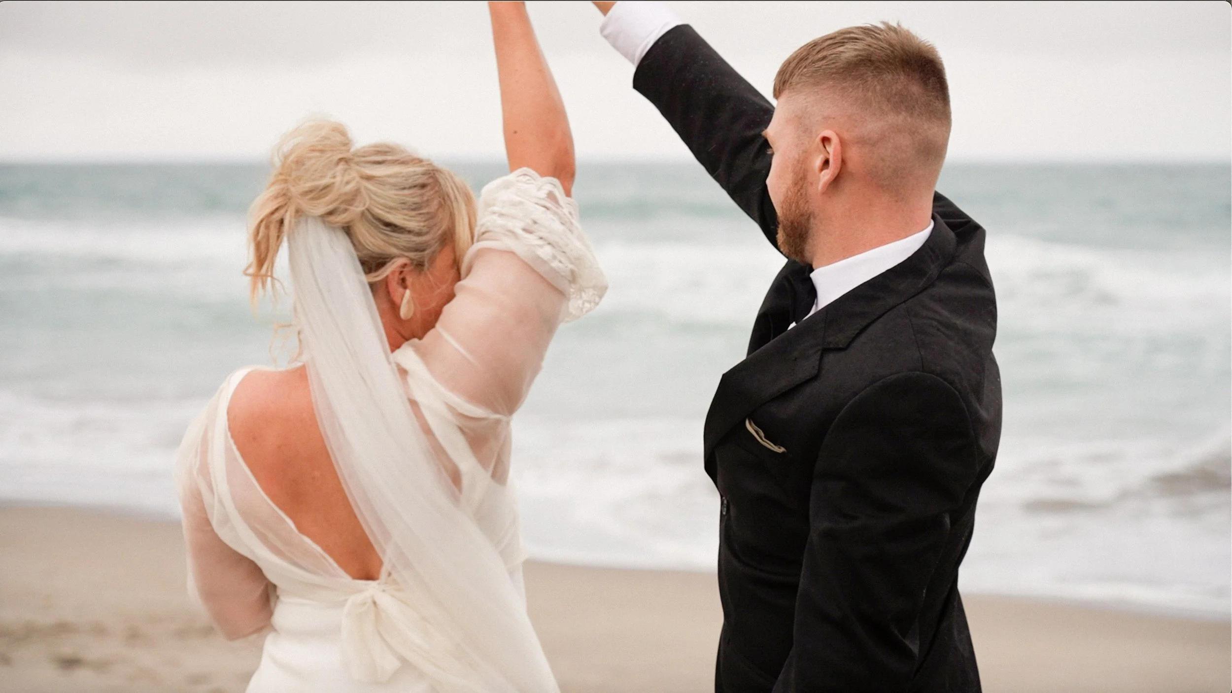 A couple on the beach during their wedding, with the woman in a white dress and veil and the man in a suit, holding hands in the air during their dance.