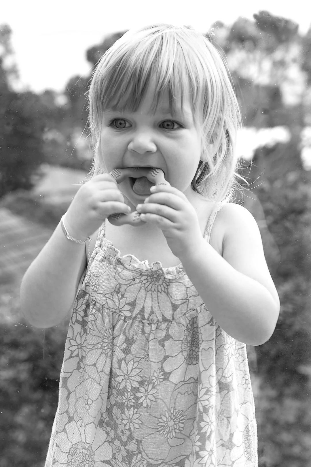 A young girl with light-colored hair, wearing a floral dress, biting into a ring-shaped snack, standing outdoors with blurred trees in the background.