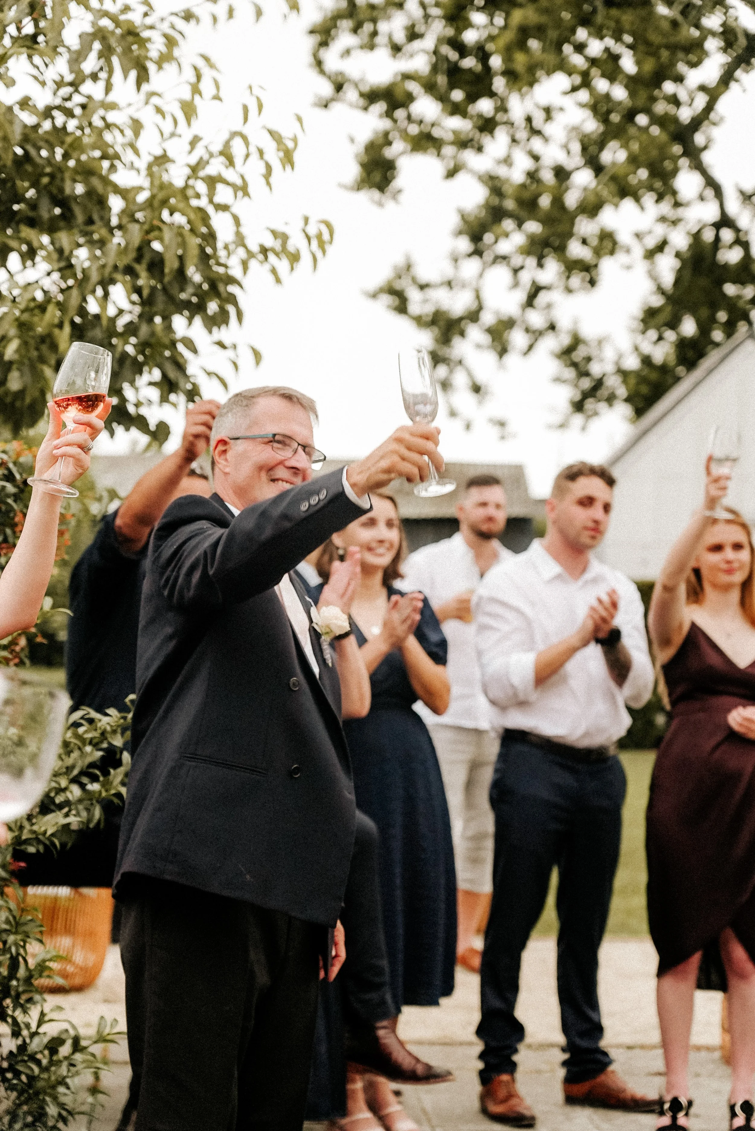 Group of people at a celebration holding up champagne glasses, smiling, and clapping outdoors in a garden setting.