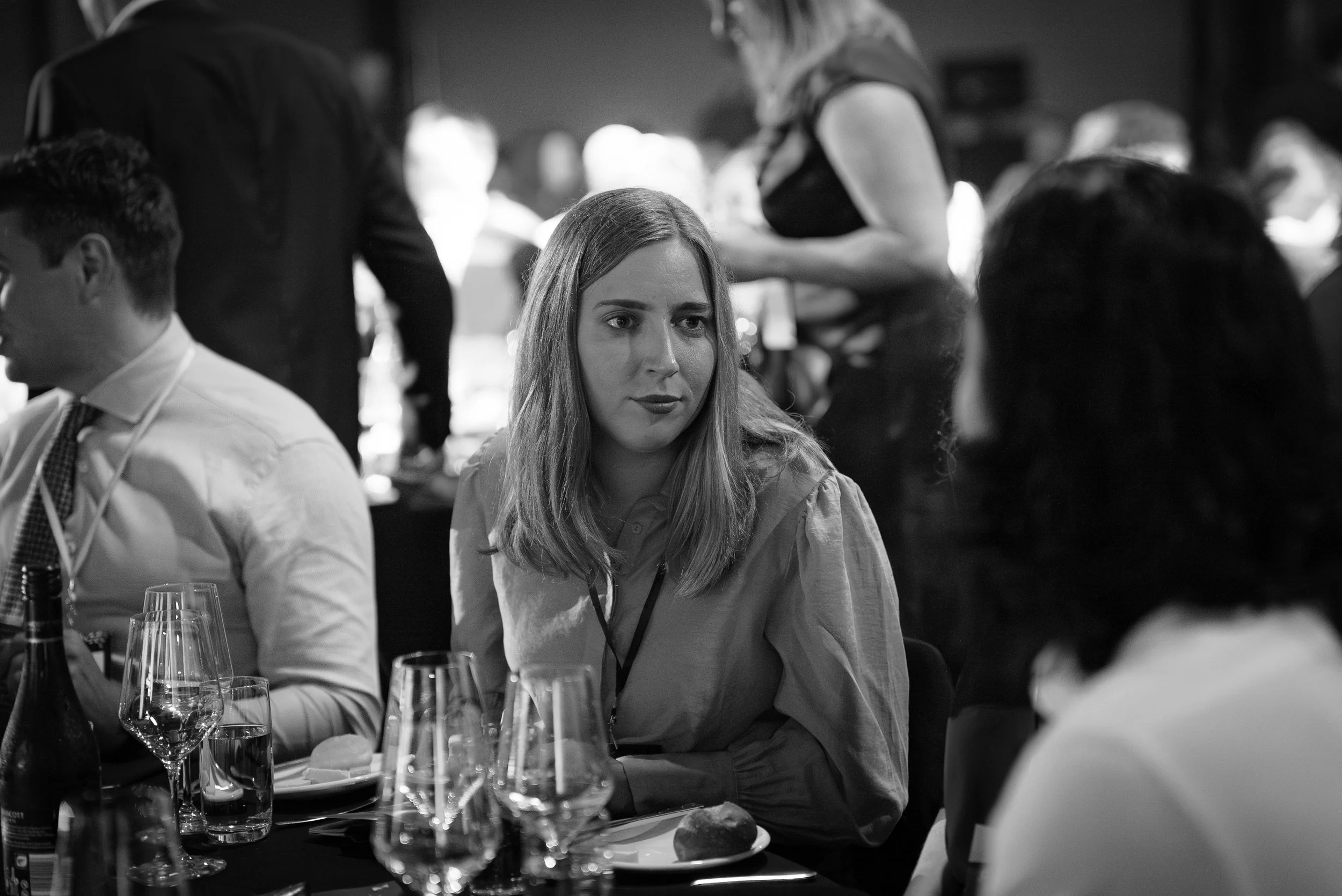 Black and white photo of a woman with shoulder-length hair sitting at a dinner table, looking at another woman with curly hair. The table has glasses, plates, and bread. Other people and waitstaff are in the background.